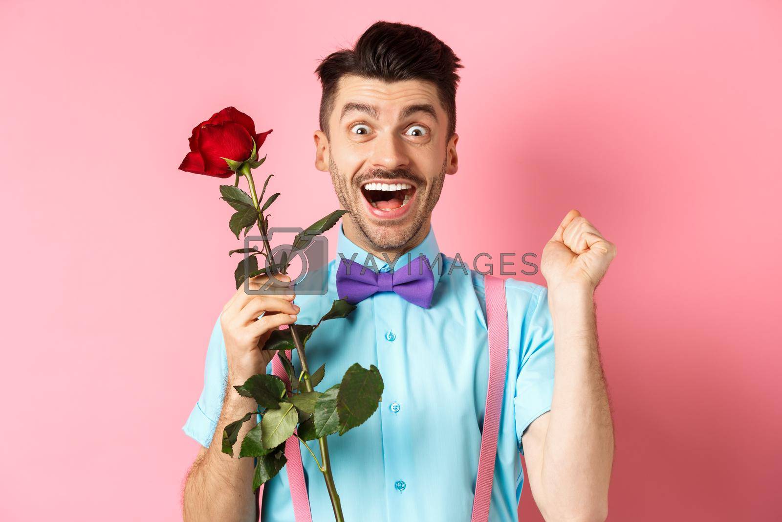 Valentines day and romance concept. Excited boyfriend jumping from happiness on romantic date, holding red rose and celebrating, standing on pink background.