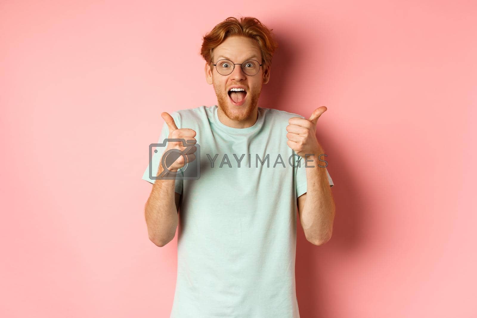 Royalty free image of Amazed redhead guy looking impressed and excited, showing thumbs up in approval, praise awesome promo offer, standing over pink background by Benzoix