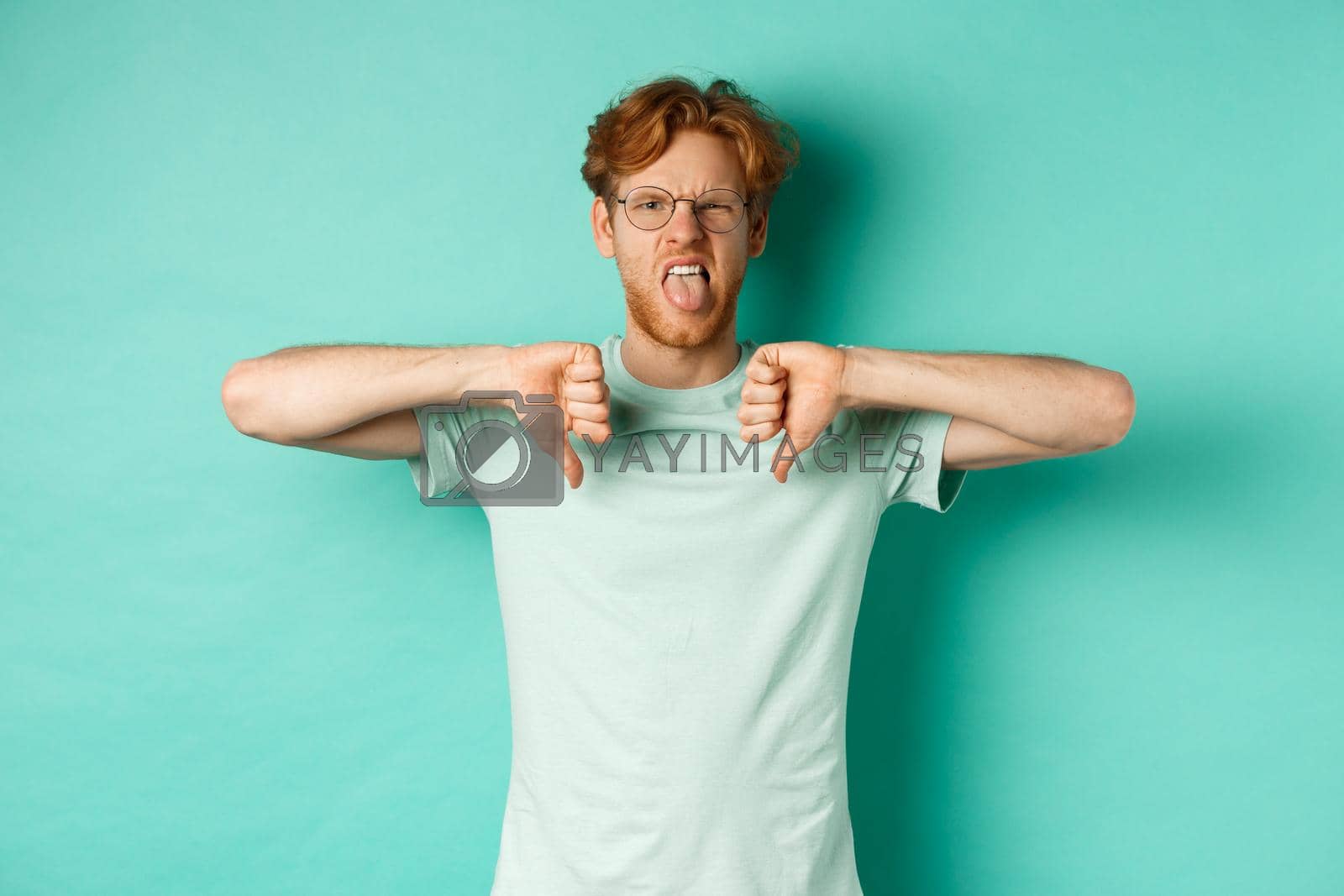 Royalty free image of Judgemental redhead male model in t-shirt and glasses, showing tongue and frowning displeased, make thumbs-down to express disappointment and dislike, turquoise background by Benzoix