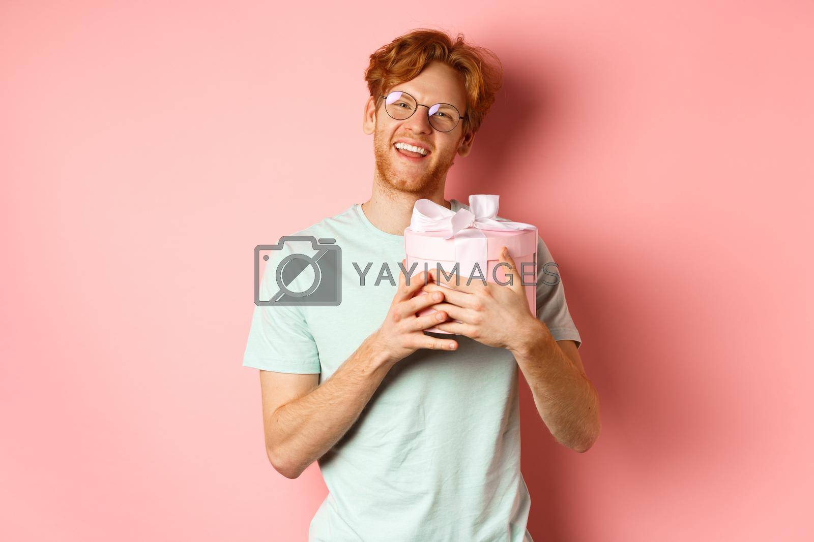 Valentines day and romance concept. Happy redhead boyfriend receive romantic gift, hugging box with present and saying thank you, smiling grateful, pink background.