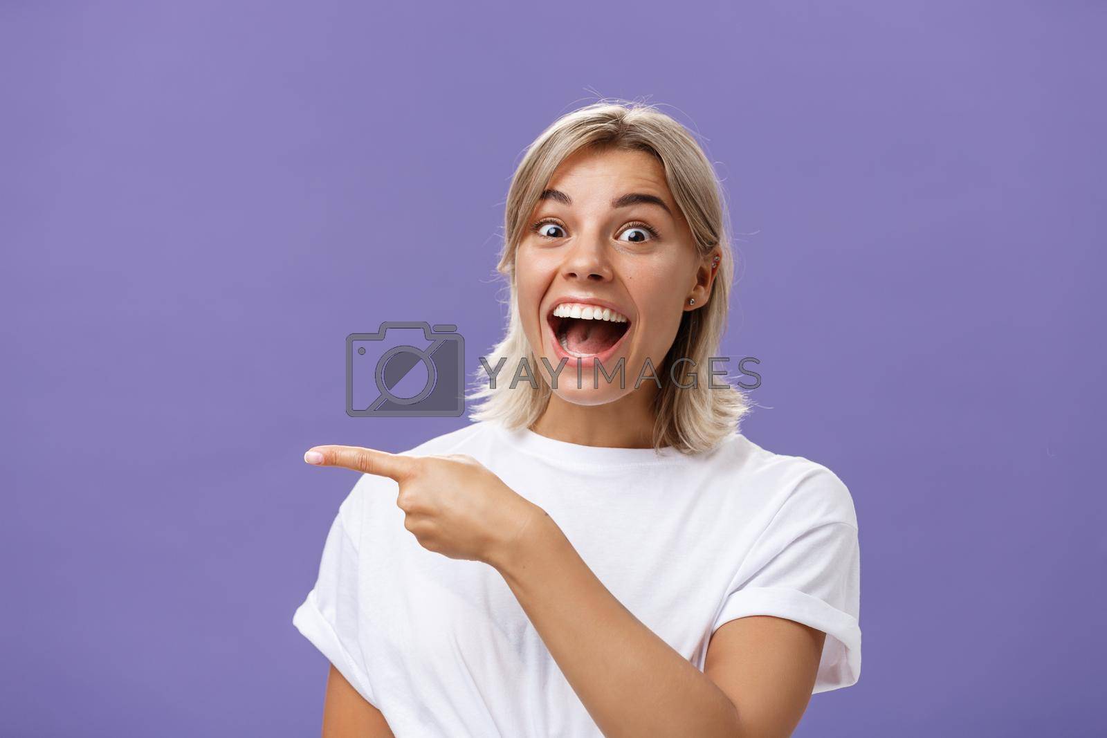 Royalty free image of Waist-up shot of joyful thrilled attractive and emotive young blonde female with tanned skin in white casual t-shirt smiling and laughing happily pointing left with excitement over purple wall by Benzoix