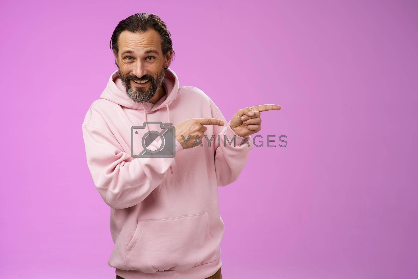 Royalty free image of Sassy confident good-looking macho man 50s in pink hoodie smiling broadly inviting join pointing right showing interesting amusing place hang out welcoming take look, standing purple background by Benzoix