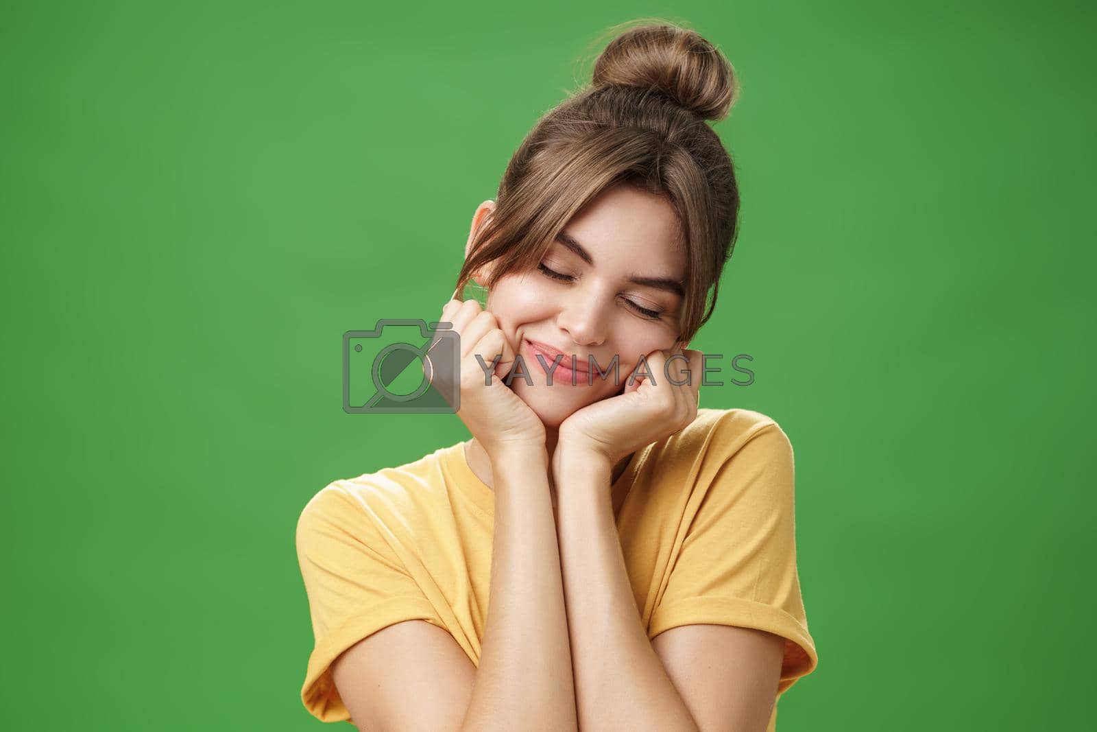 Royalty free image of Tender sensual timid european girl in yellow t-shirt leaning head on shoulder touching cheeks with hands closing eyes and smiling with soft grin feeling nostalgic and romantic over green background by Benzoix