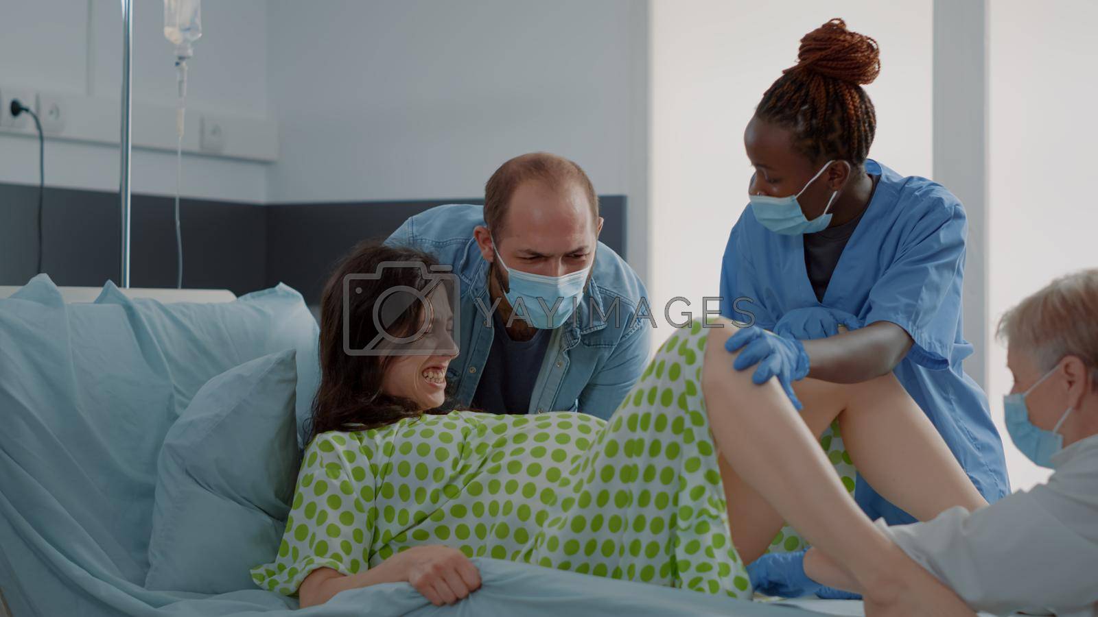 Multi ethnic people delivering child in hospital ward at maternity. Pregnant woman in labor pushing, giving birth while husband, obstetrics doctor and african american nurse helping patient