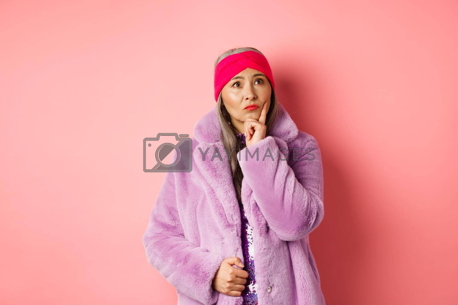 Royalty free image of Funky old asian woman looking thoughtful and perplexed, standing in fake fur coat and thinking, making decision, standing on pink background by Benzoix