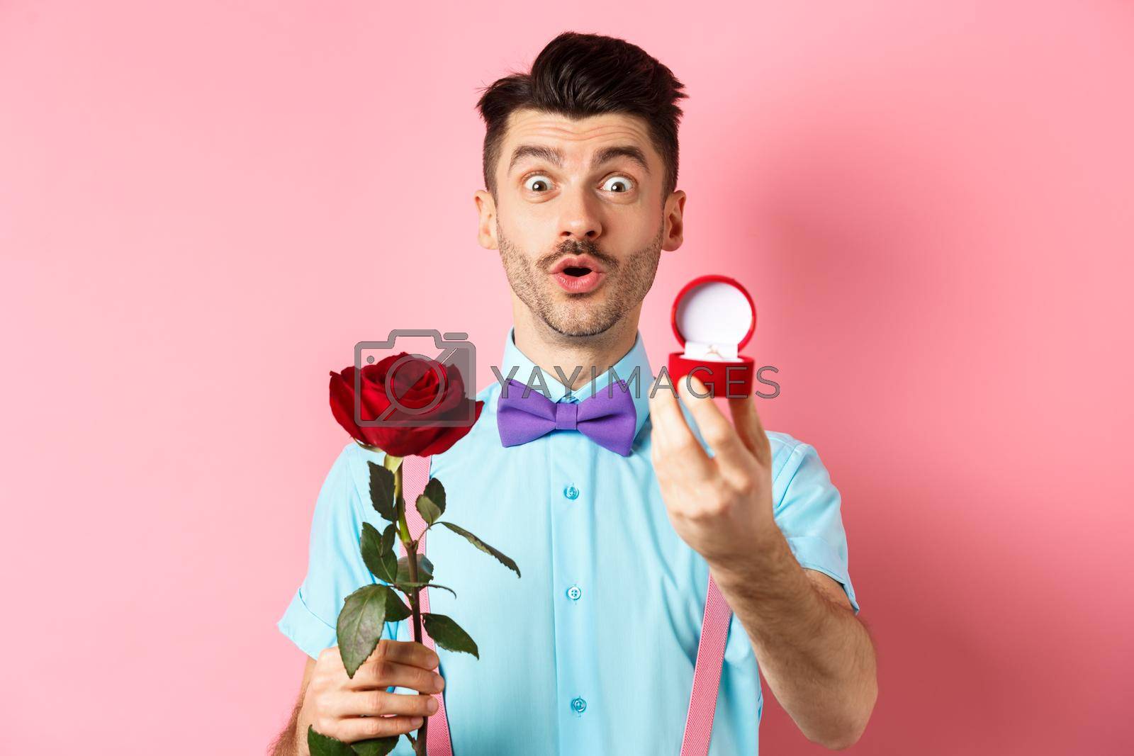 Royalty free image of Valentines day. Funny man with moustache and bow-tie making proposal, showing engagement ring and propose with red rose, standing over pink background by Benzoix