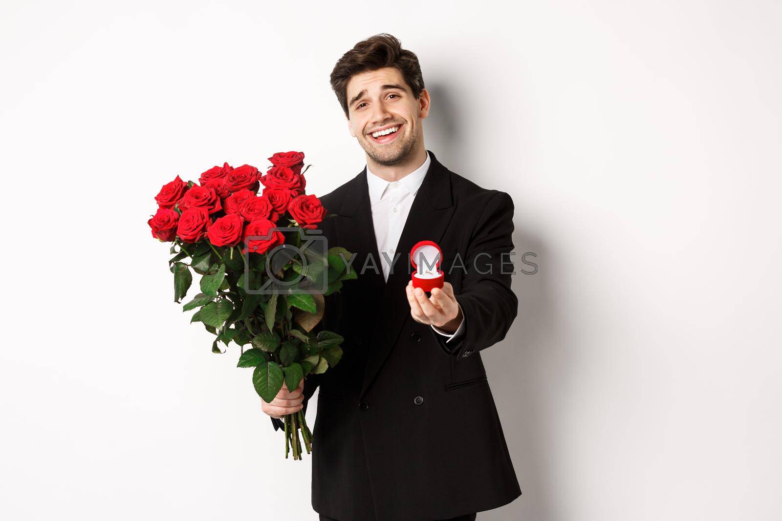 Royalty free image of Handsome smiling man in black suit, holding roses and engagement ring, making a proposal to marry him, standing against white background by Benzoix