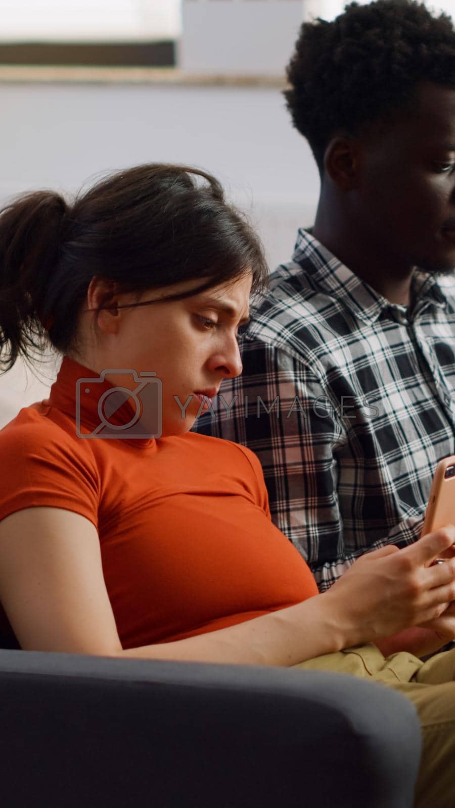 Modern interracial people holding smartphones on couch while sitting together in living room. Young mixed race couple using devices and technology for entertainment as lifestyle
