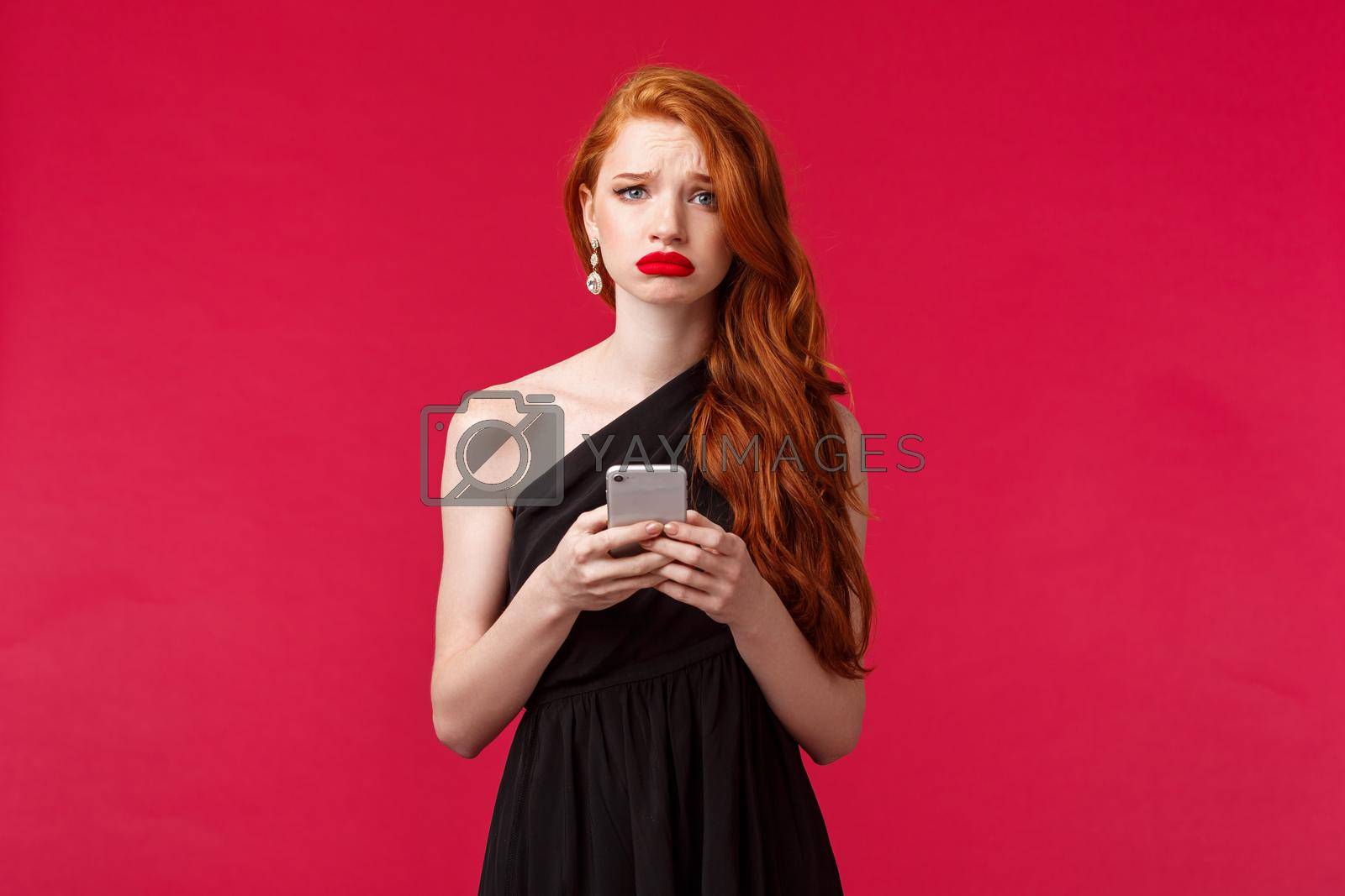 Royalty free image of Portrait of gloomy upset gorgeous redhead girl in black dress, sobbing and grimacing unhappy, holding mobile phone, receive disappointing news via message, complaining, red background by Benzoix
