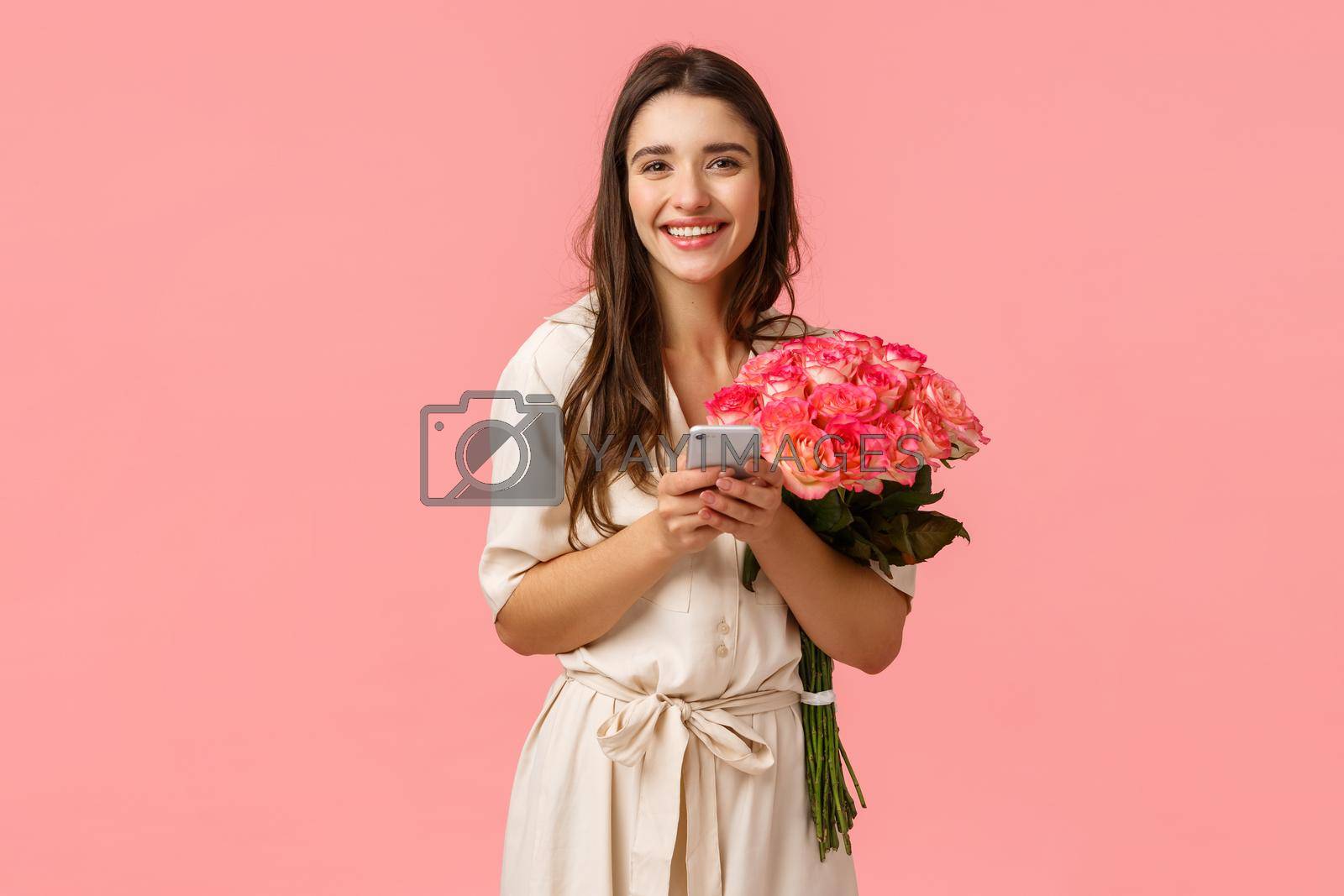 Technology, romance and happiness concept. Tender young cheerful smiling woman with beautiful flowers, holding smartphone, answer on congratulations b-day, chatting on birthday, pink background.