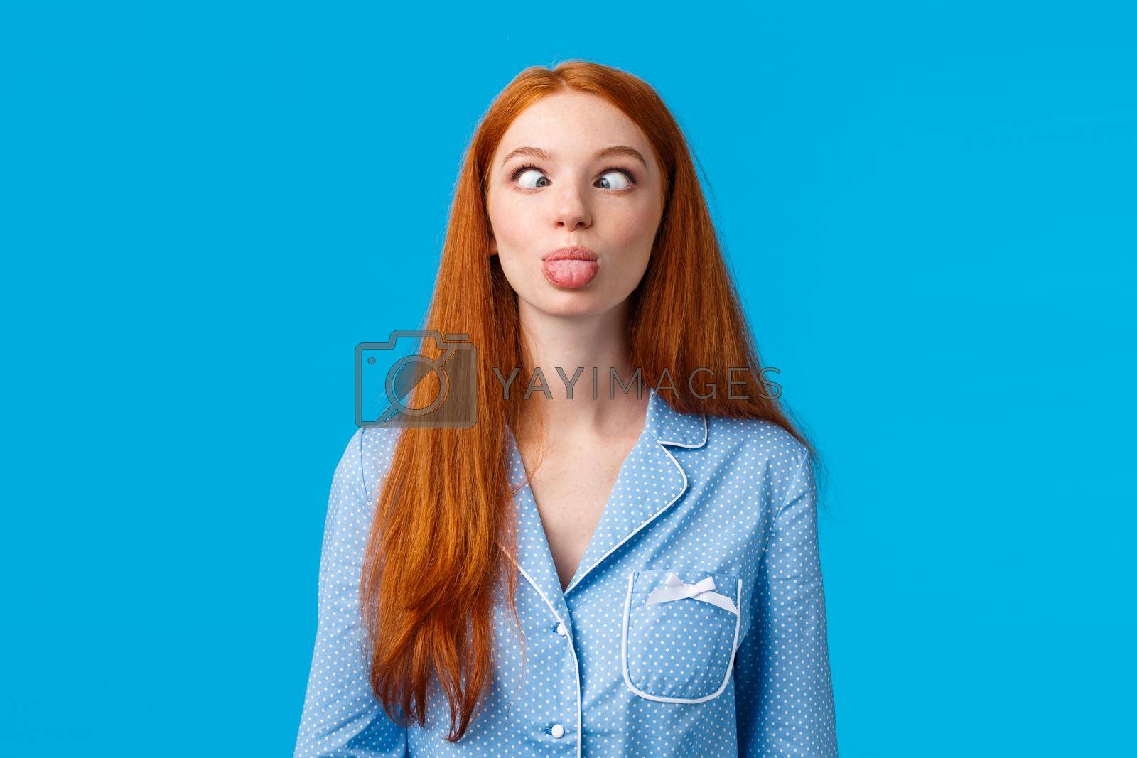 Royalty free image of Be funny and silly. Redhead carefree and enthusiastic caucasian girl squinting, making goofy expression, showing tongue, standing blue background in nightwear, fool around by Benzoix