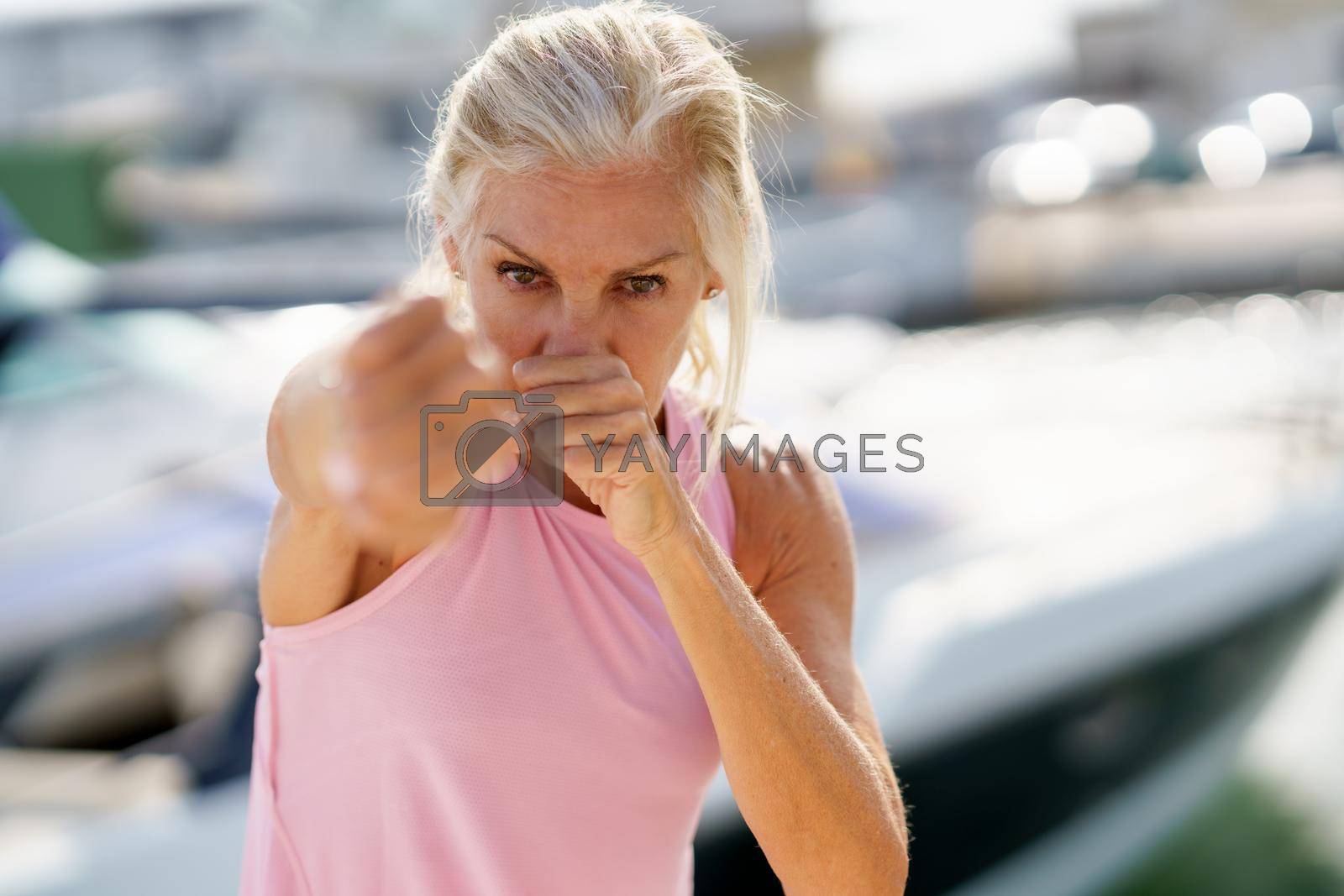 Mature woman doing sport in a coastal port. Arm training throwing boxing punches. Female doing shadow boxing outdoors.