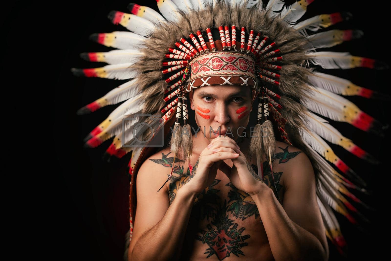 Royalty free image of a white man in a headdress of the indigenous peoples of North America with a naked torso on a dark background by Rotozey