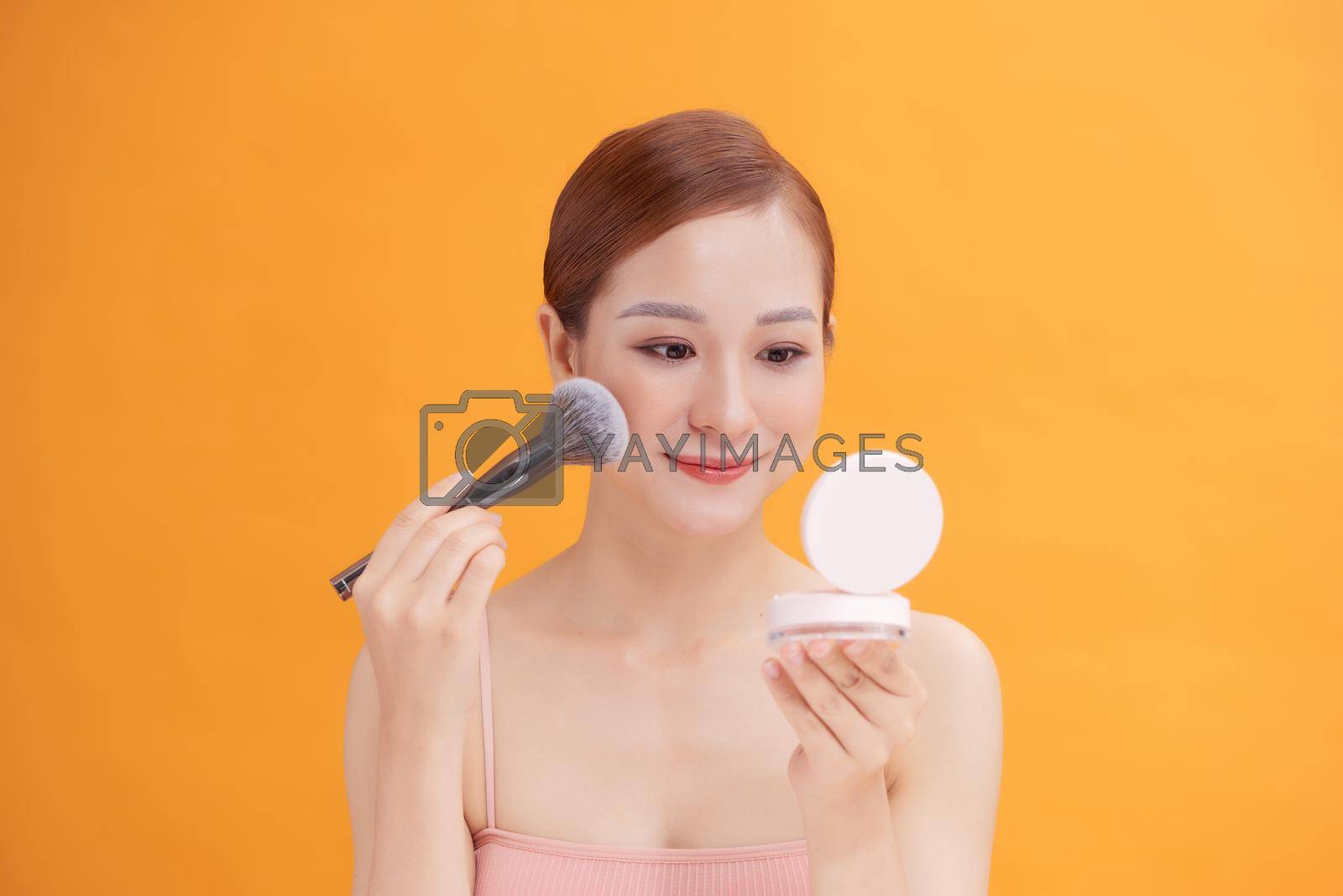 Beautiful woman applying blusher to her cheek with a large cosmetics brush while holding a compact mirror in front of her