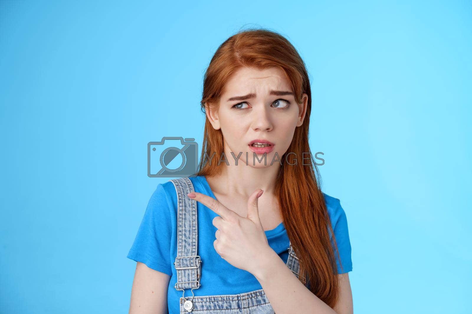 Royalty free image of Stupified redhead hesitant girl show aversion, pointing looking left reluctant, full disbelief, stare with dismay uncertain about product, disliking strange weird guy standing blue background by Benzoix
