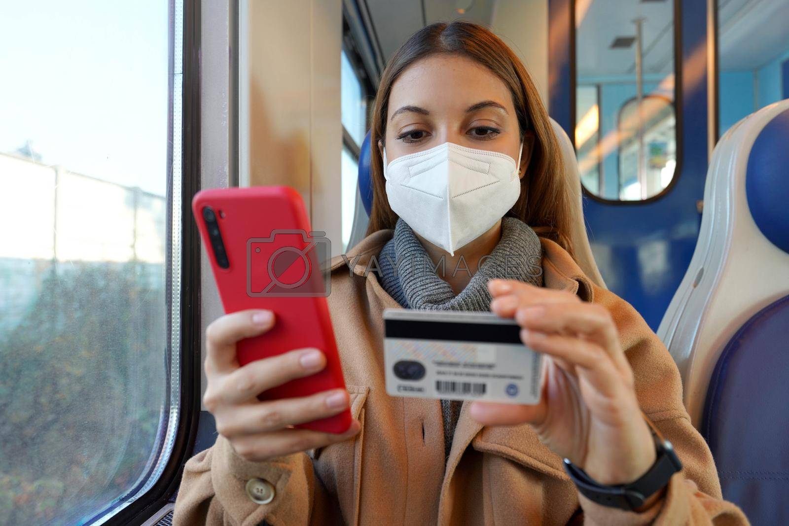 Portrait of woman with FFP2 KN95 protective mask traveling with train paying with her credit card on smart phone for shopping online