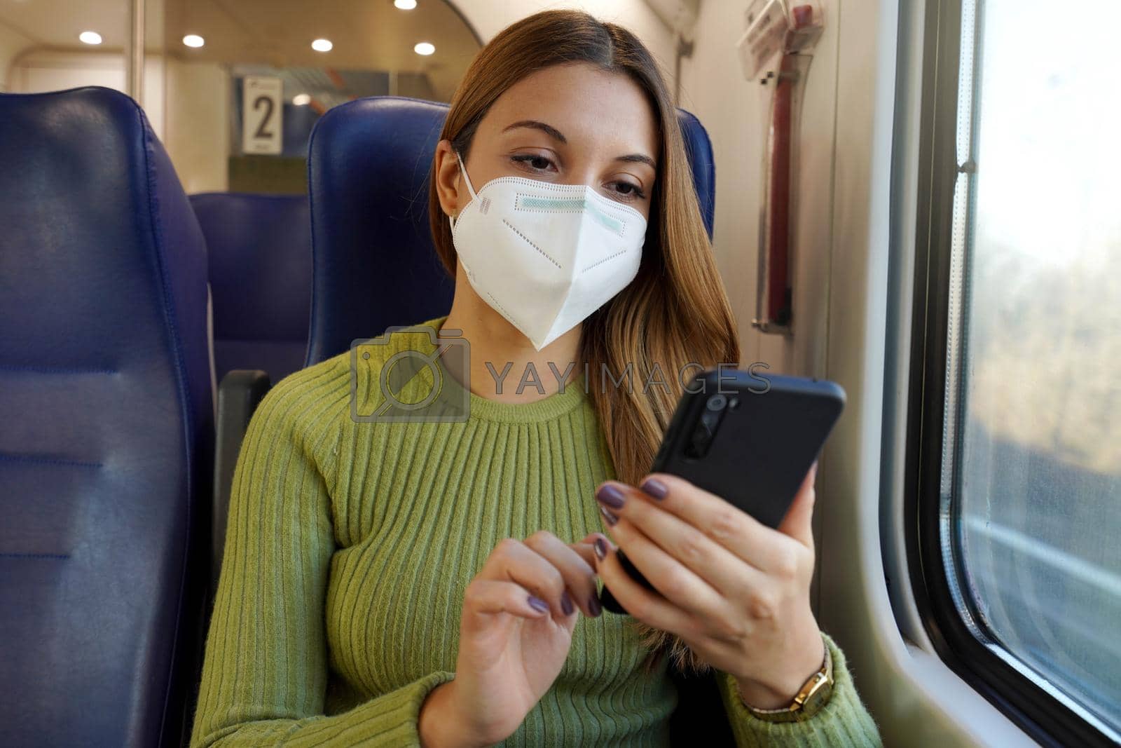 Relaxed woman on train wearing medical face mask using smart phone app. Commuter with protective mask traveling sitting in business class texting on mobile phone. Travel safely on public transport.