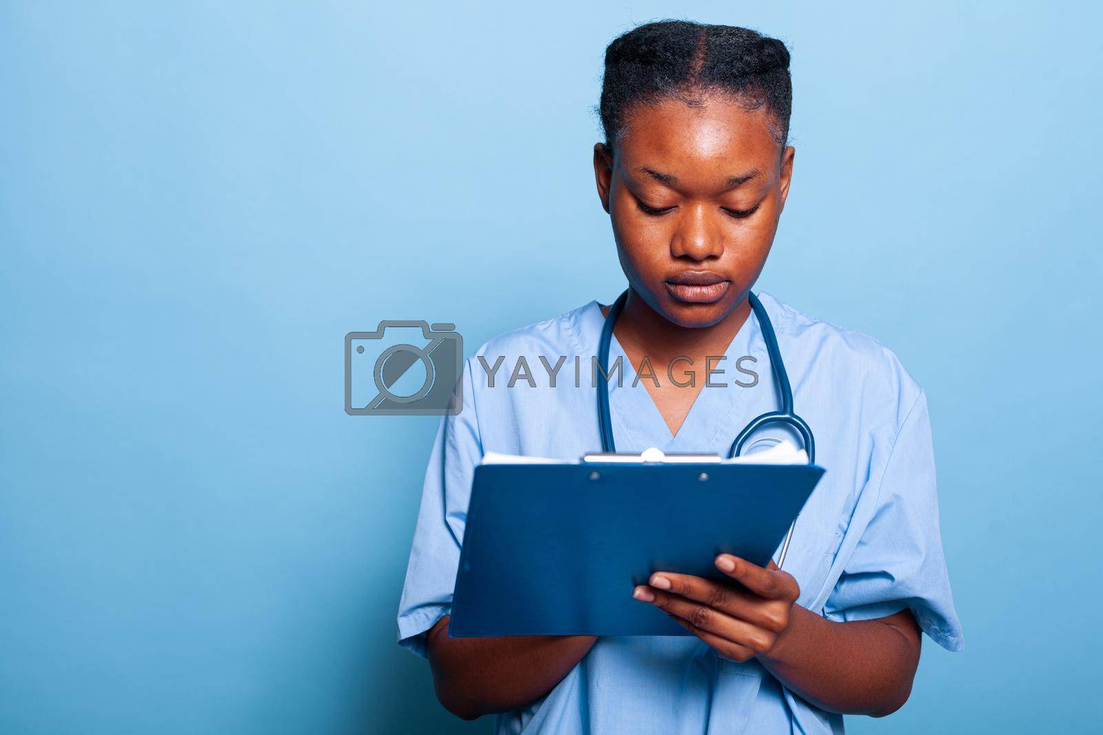 African american practitioner assistant writing healthcare treatment on clipboard working at sickness expertise in studio with blue background. Therapist nurse in medical uniform. Medicine concept