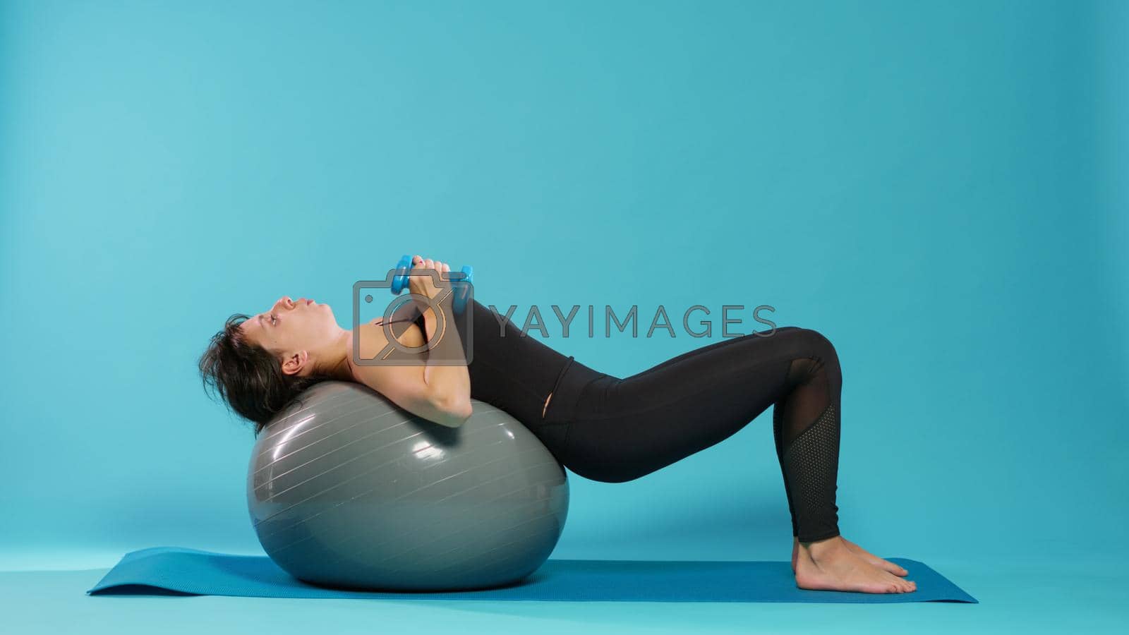 Active woman lifting dumbbells on fitness toning ball, doing physical exercise to train muscles in studio. Young adult using workout equipment and weights to practice sport and wellbeing.