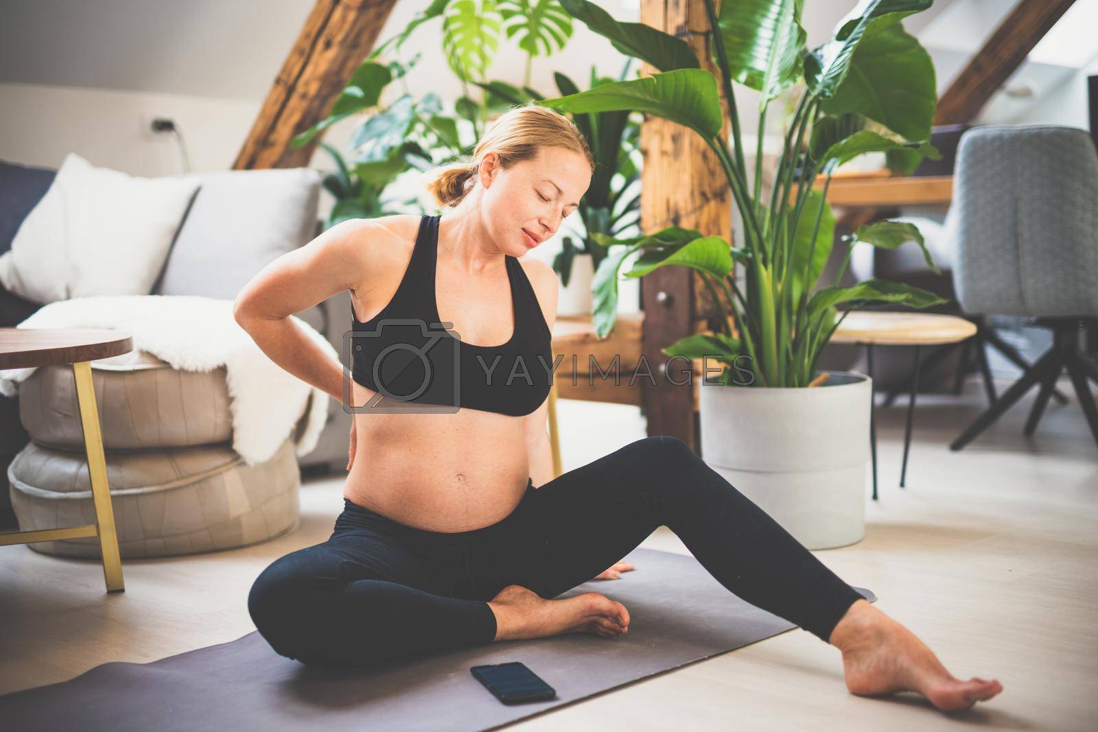 Young beautiful pregnant woman training yoga and stretching, feeling backpain at home in her living room. Motherhood, pregnancy, healthy lifestyle and yoga concept