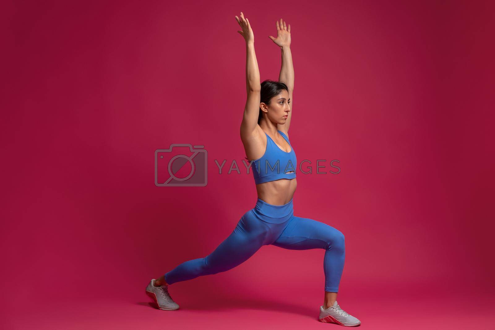Sporty young woman performing Virabhadrasana, lunging standing asana with arms stretched straight upwards. Studio portrait on maroon background. Active lifestyle and yoga concept