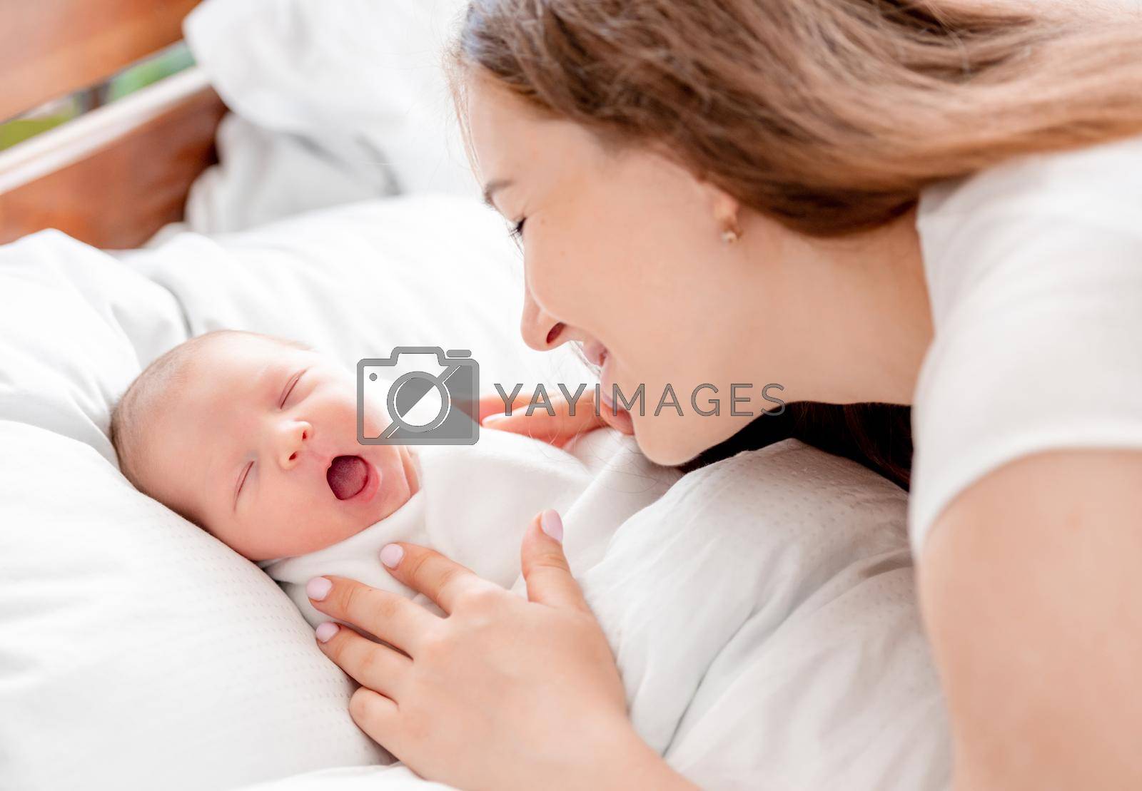 Young mother looking at her sleeping with opened mouth newborn daughter swaddled in white sheets at home and smiling with tenderness. Portrait of girl mom with her infant child napping in the bed