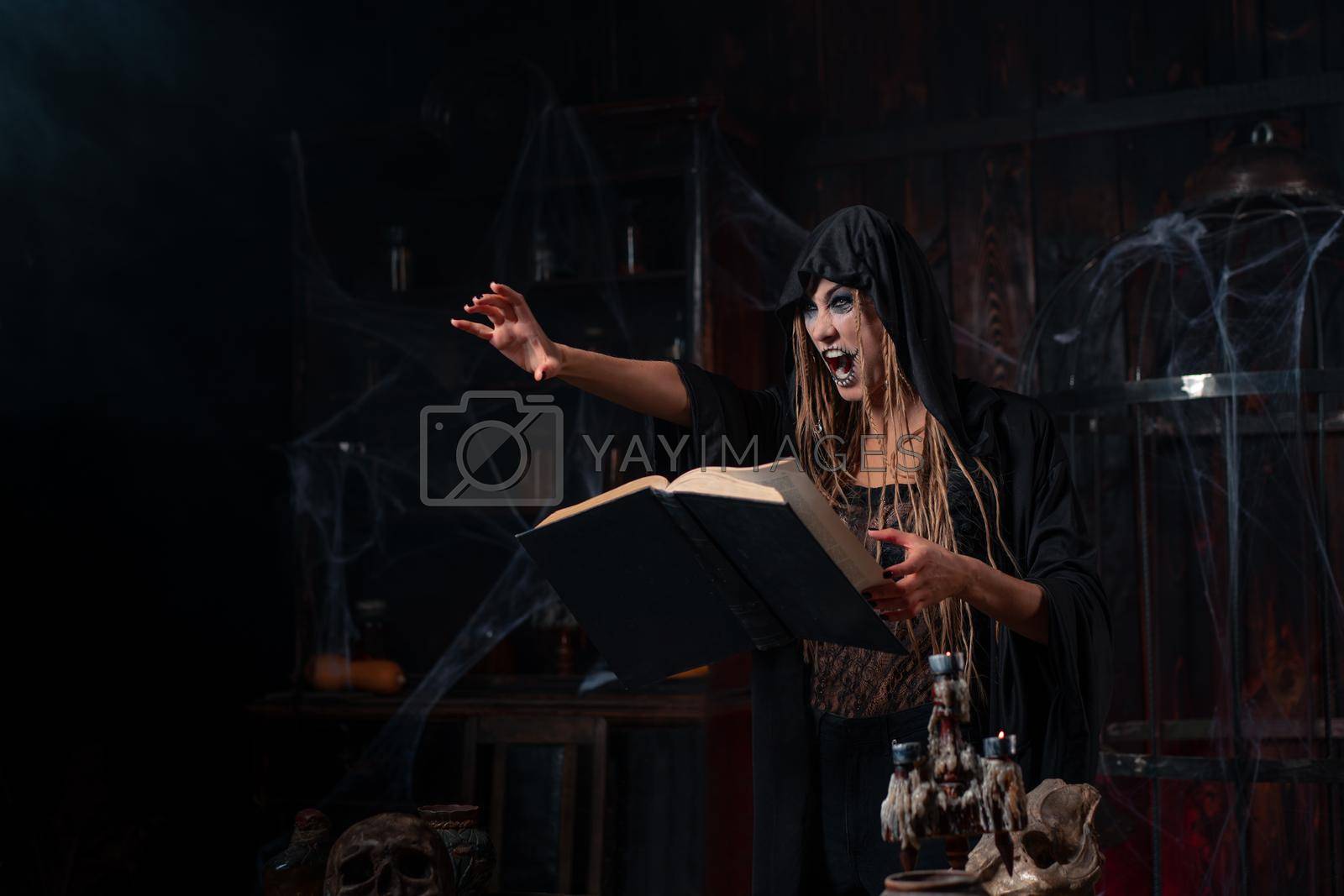 Royalty free image of Halloween concept. Witch dressed black hood with dreadlocks standing dark dungeon room use magic book for conjuring magic spell by andreonegin