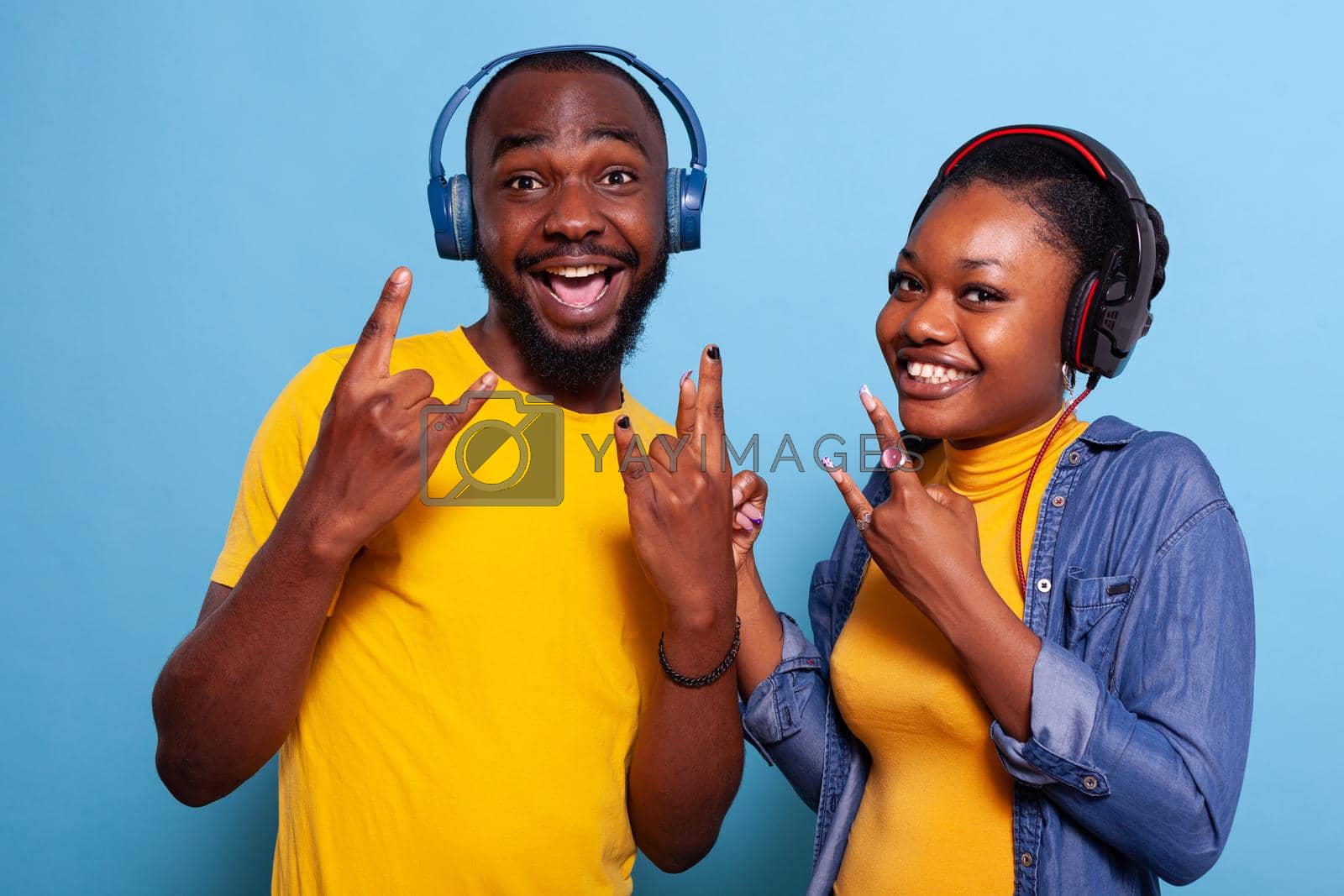 Happy people showing rock symbol with fingers on camera, listening to music on headphones. Playful couple enjoying song and rhythm on headset, doing hands gesture and smiling.