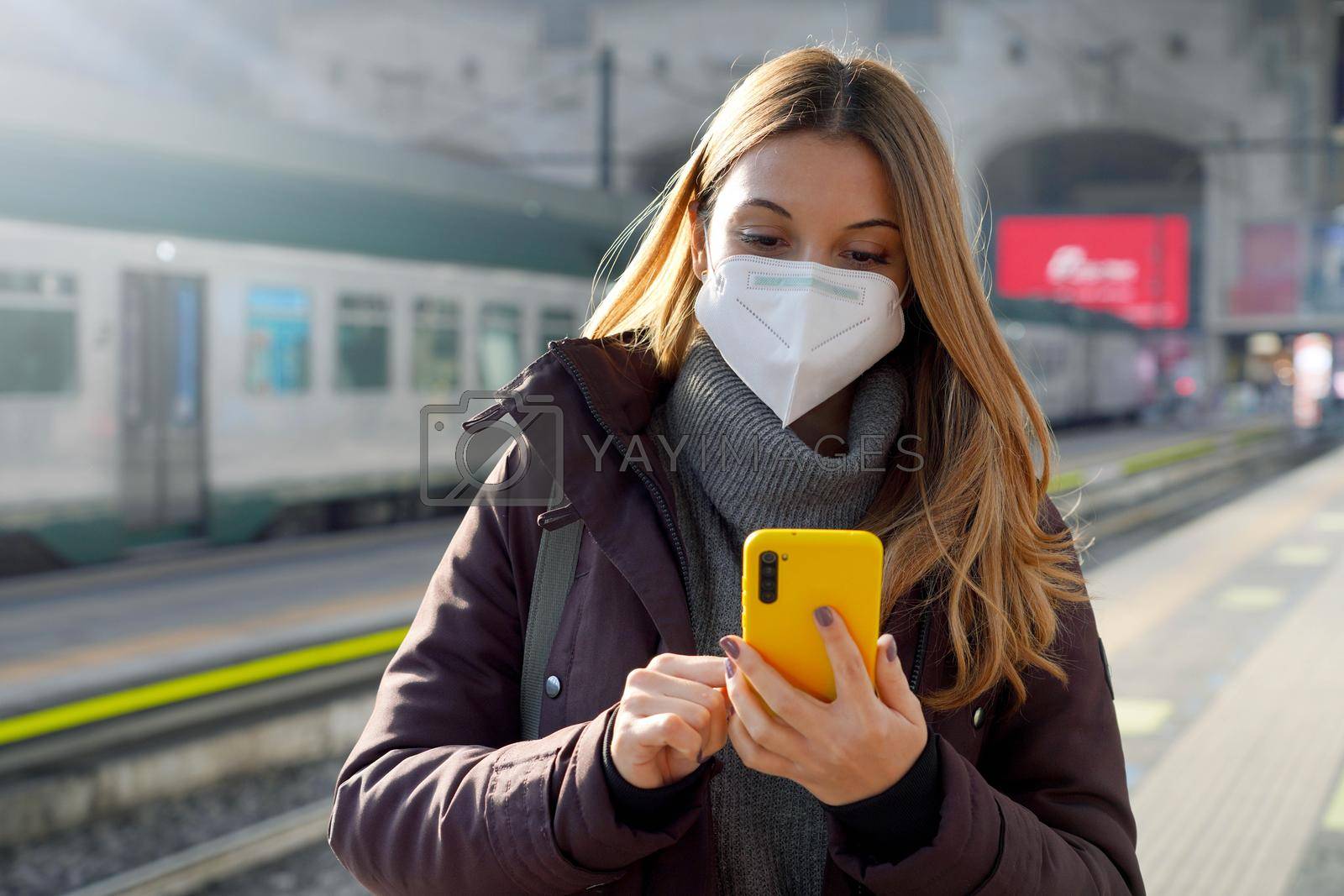 Portrait of a girl in a protective mask standing on the platform of train station with a mobile phone buying ticket online