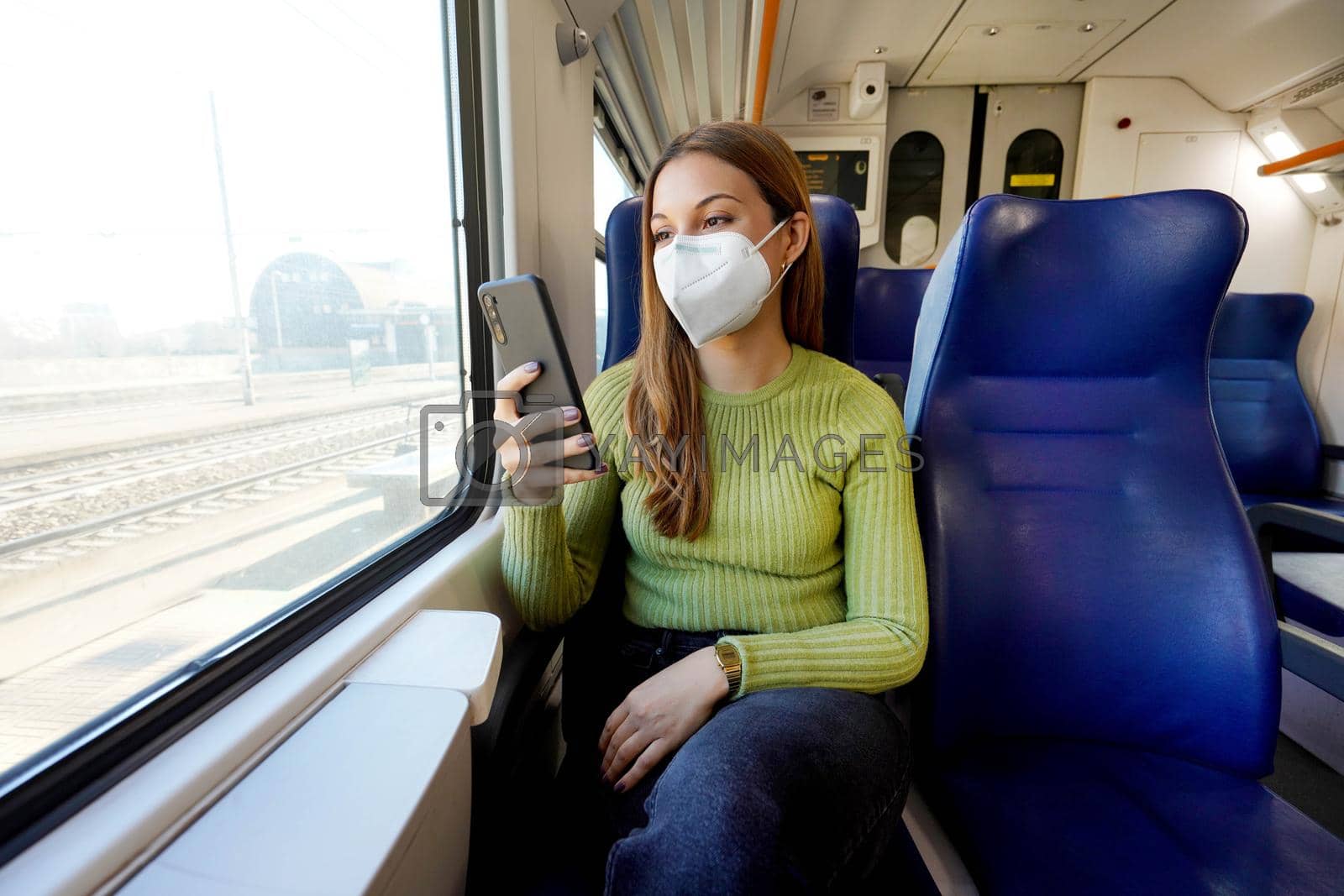 Young woman wearing medical mask relaxing in train seat while using smartphone app. Business woman enjoying view texting on mobile phone. Travel safety lifestyle.
