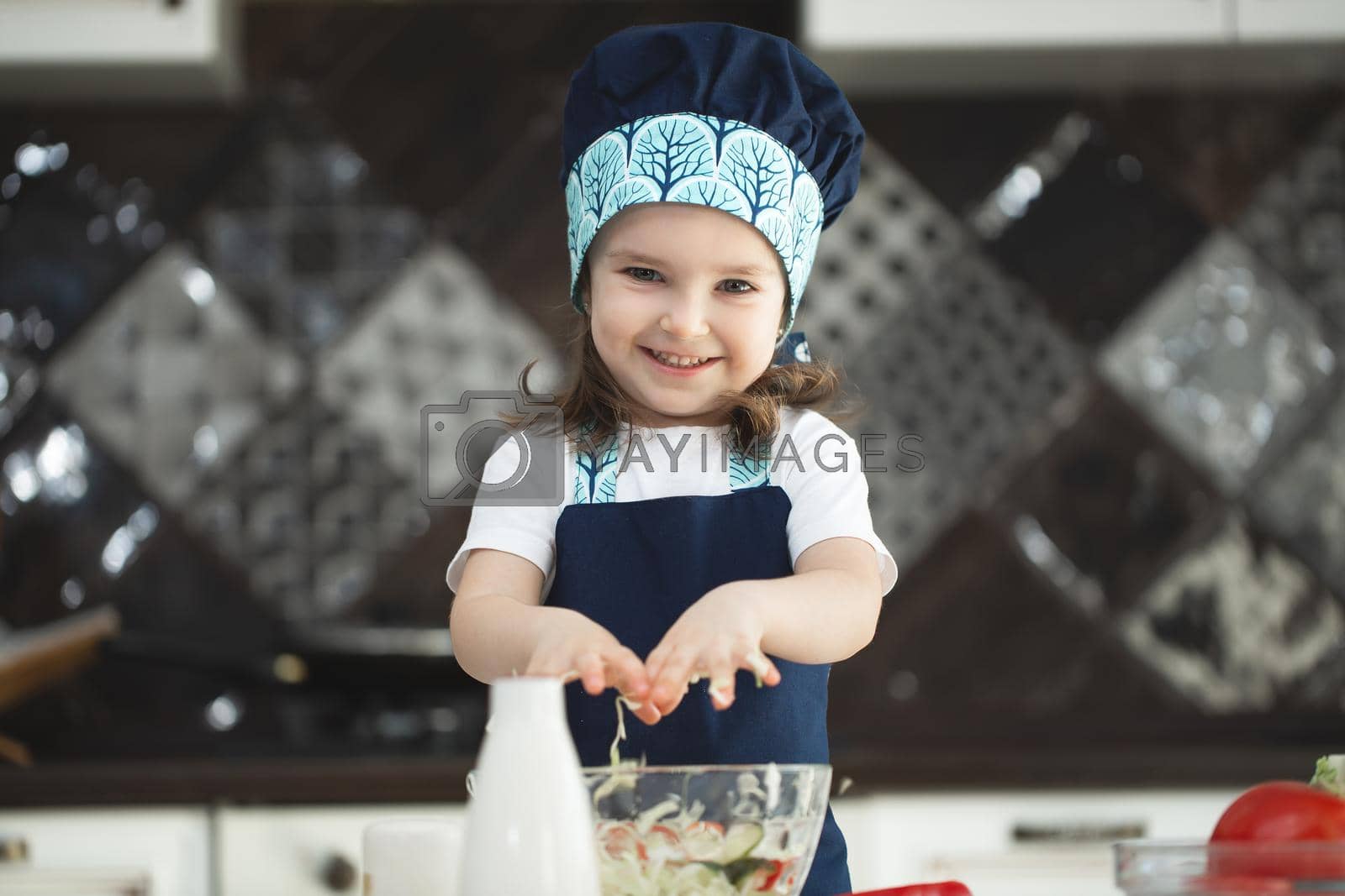 Royalty free image of A child in an apron and a Chef's hat is stirring a vegetable salad in the kitchen and looking at the camera by StudioPeace