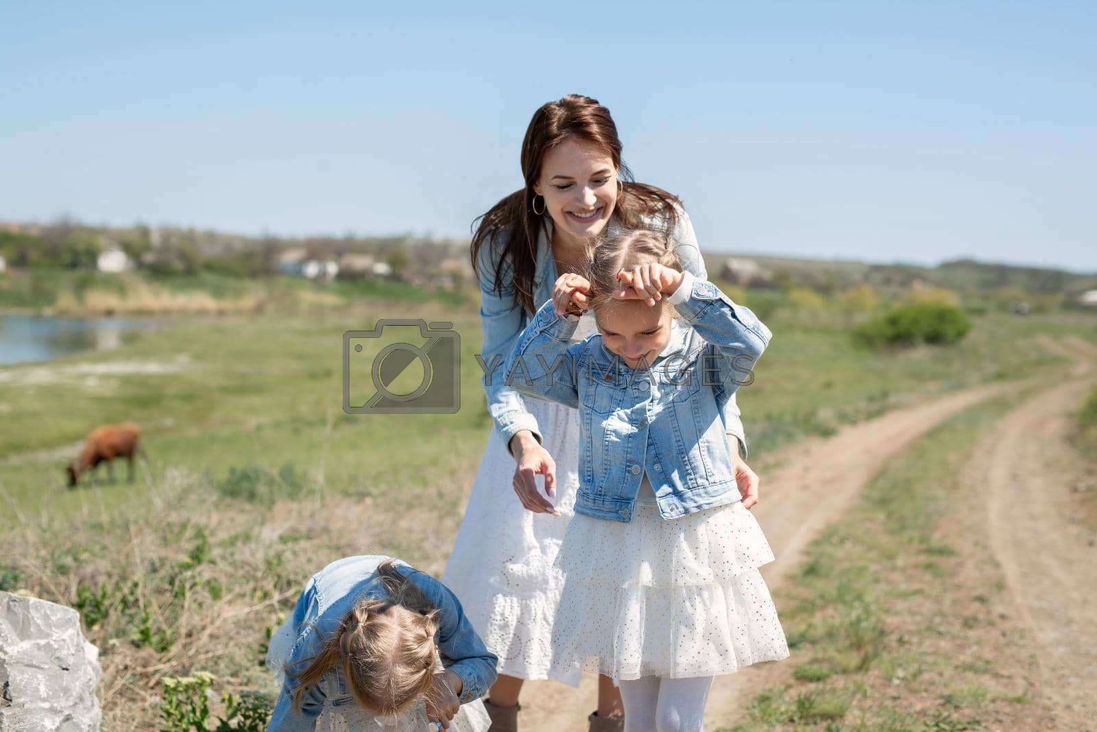 Royalty free image of Mother with her twin daughters walks along a dirt road in a village near cows. Mother and daughters pretend to be cows and have fun by StudioPeace