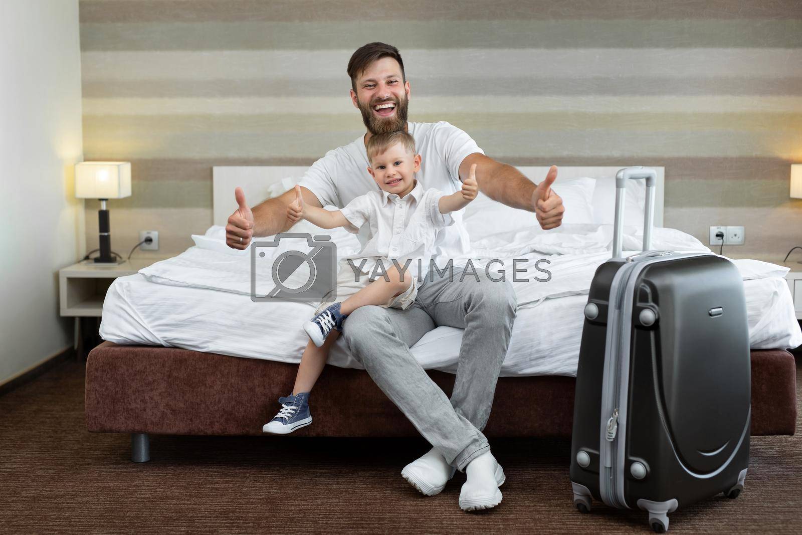 Royalty free image of A young father and son sit on a bed in a hotel with a suitcase and laugh by StudioPeace