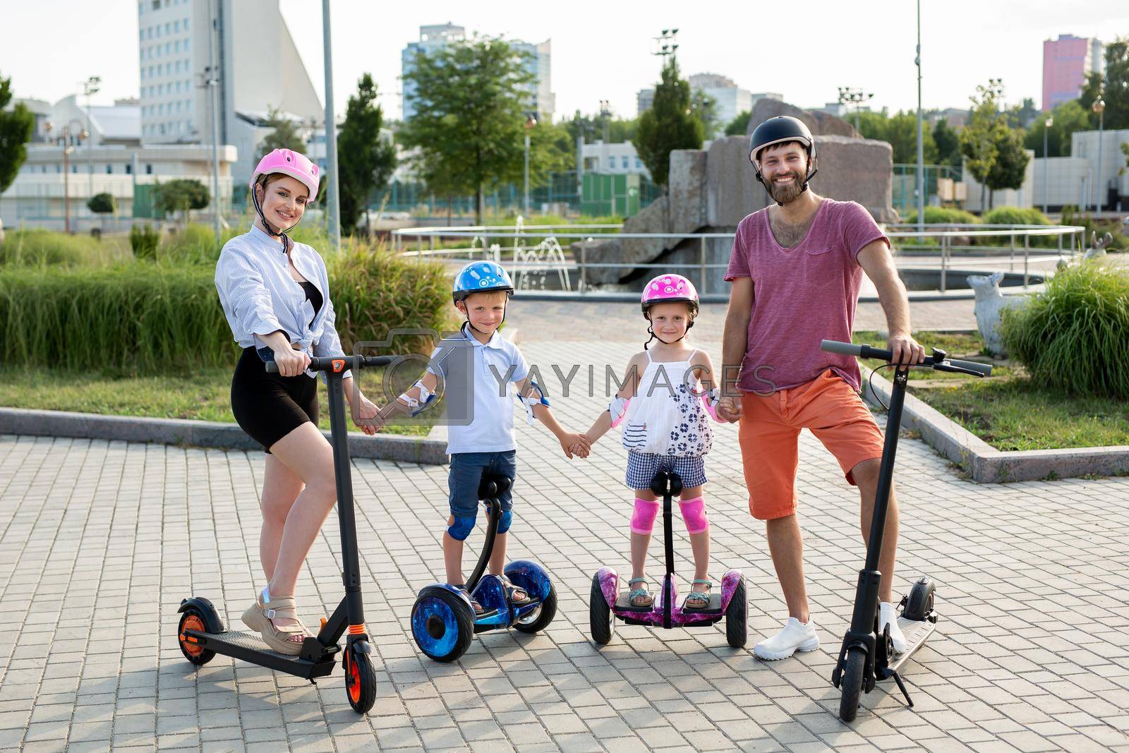 Father, mother, daughter and son in helmets stand in the park on electric scooters. Active lifestyle.