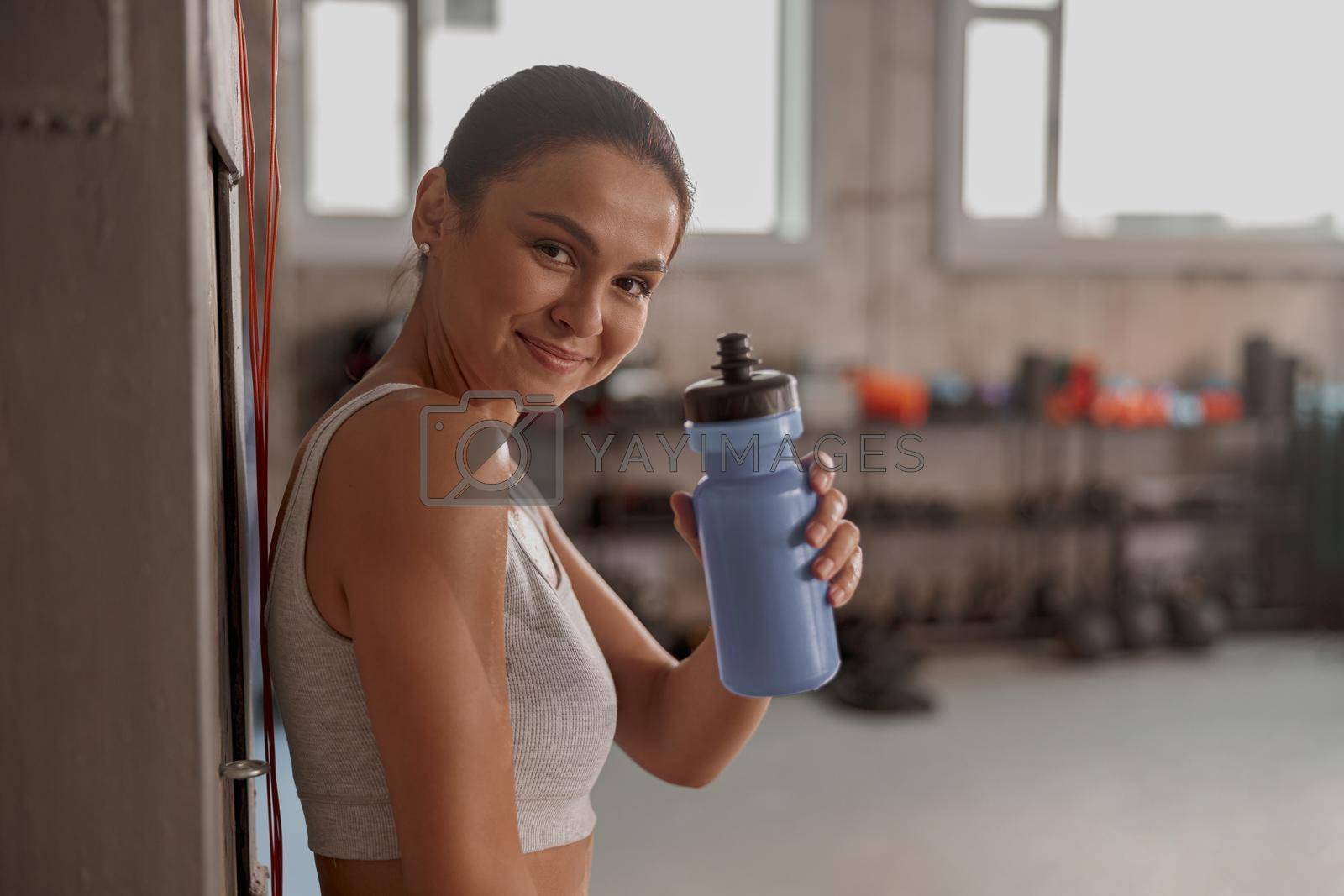 Portrait of pretty female leaning against wall at gym, holding a bottle of water and looking at camera