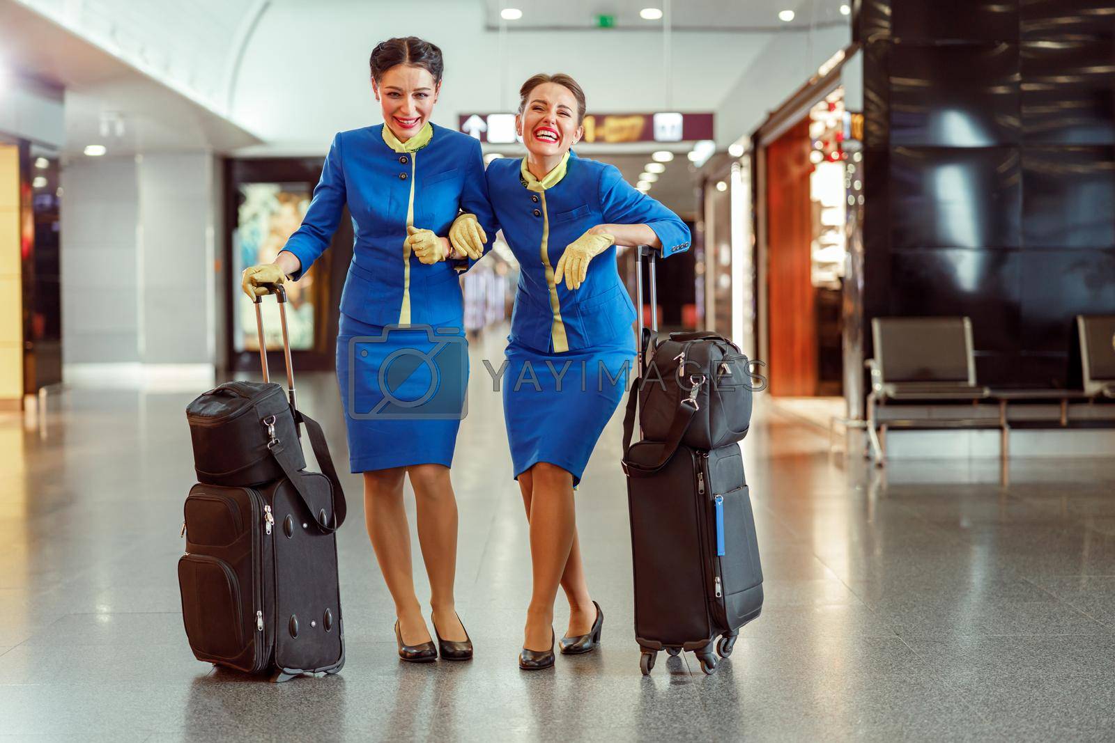 Joyful women flight attendants in aviation air hostess uniform looking at camera and laughing while standing near trolley luggage bags