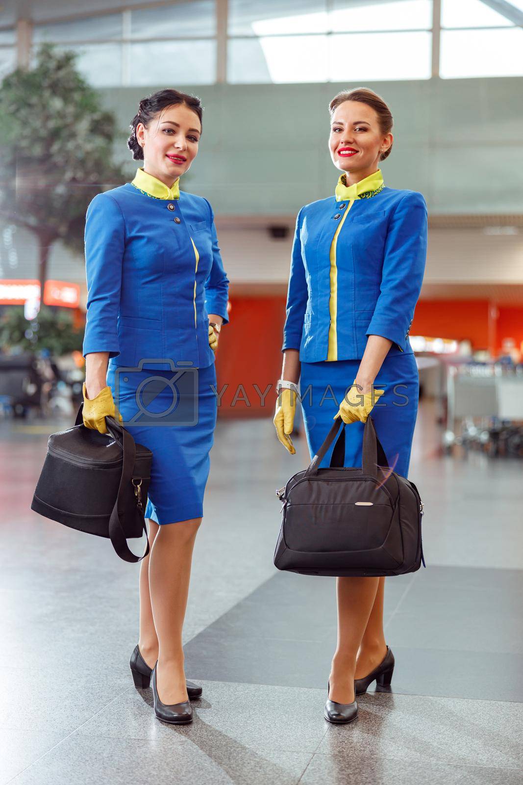 Full length of women flight attendants in aviation air hostess uniform looking at camera and smiling while holding bags