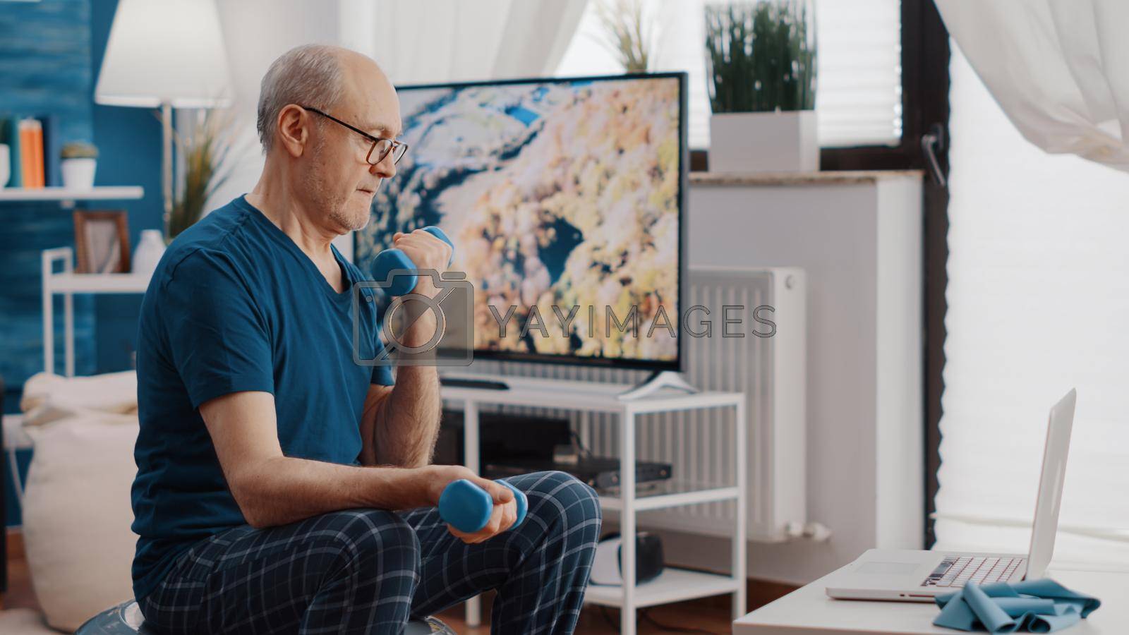 Man lifting dumbbells while sitting on fitness toning ball an looking at online video of workout on laptop. Retired adult doing exercise with weights and watching training lesson on device