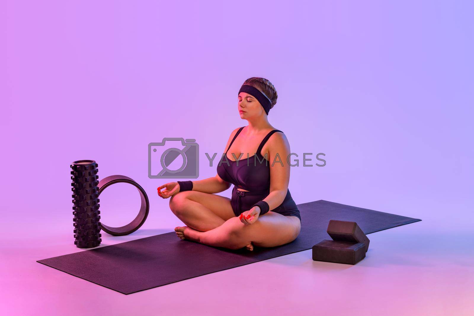 Portrait of fat young woman practicing yoga indoor. Beautiful size plus model practice asana in class. Calmness and relax on pink background