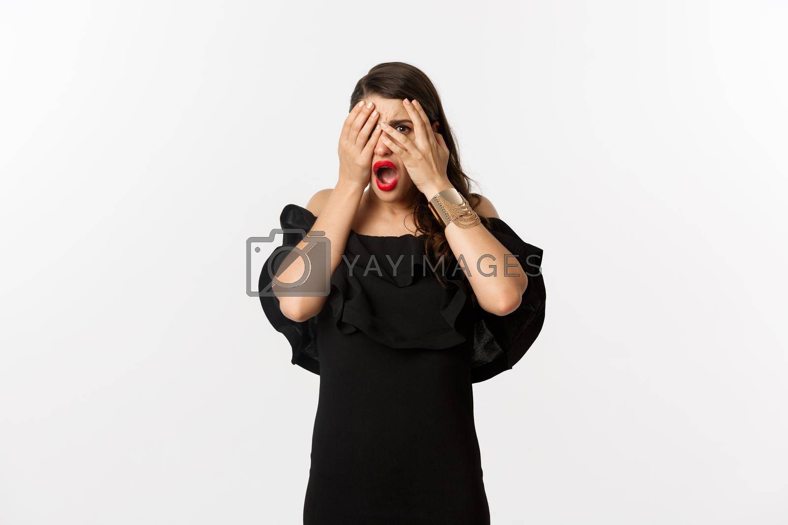 Fashion and beauty. Shocked young woman in black dress covering eyes, peeking through fingers at something embarrassing, cringe, standing over white background.