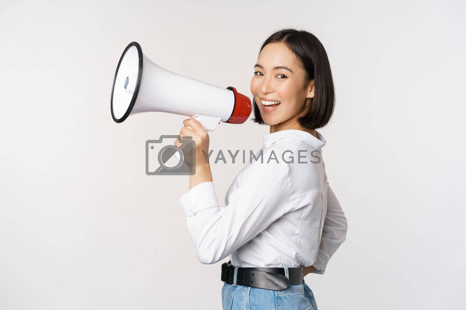Royalty free image of Beautiful young asian woman talking in megaphone, screams in speakerphone and smiling, making announcement, shout out information, standing over white background by Benzoix