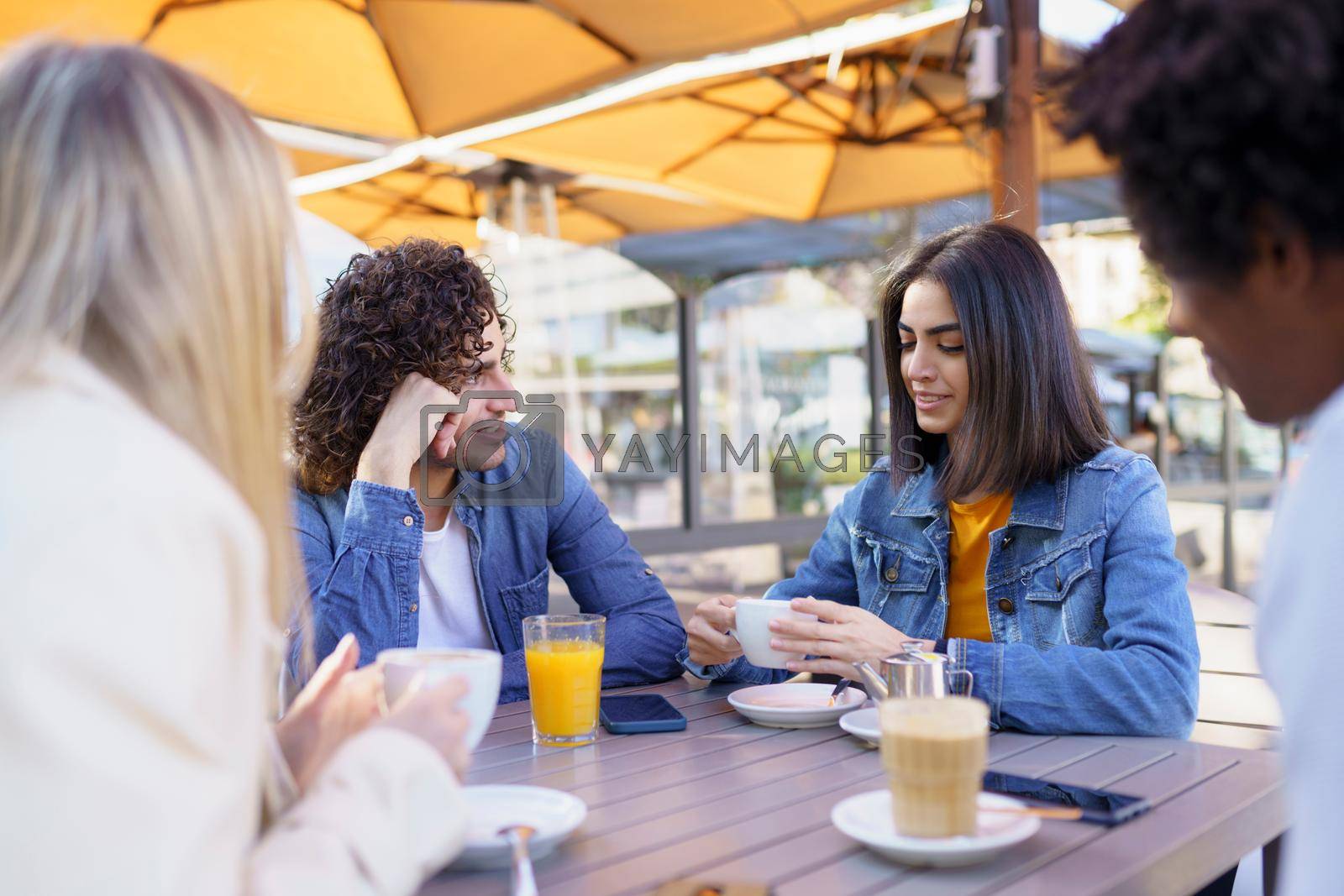 Group of smiling multiracial friends gathering around table in outdoors cafe and chatting while enjoying meeting at summer weekend