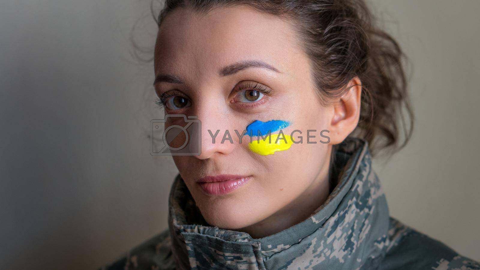 Royalty free image of Indoor portrait of young girl with blue and yellow ukrainian flag on her cheek wearing military uniform, mandatory conscription in Ukraine, equality concepts by balinska_lv