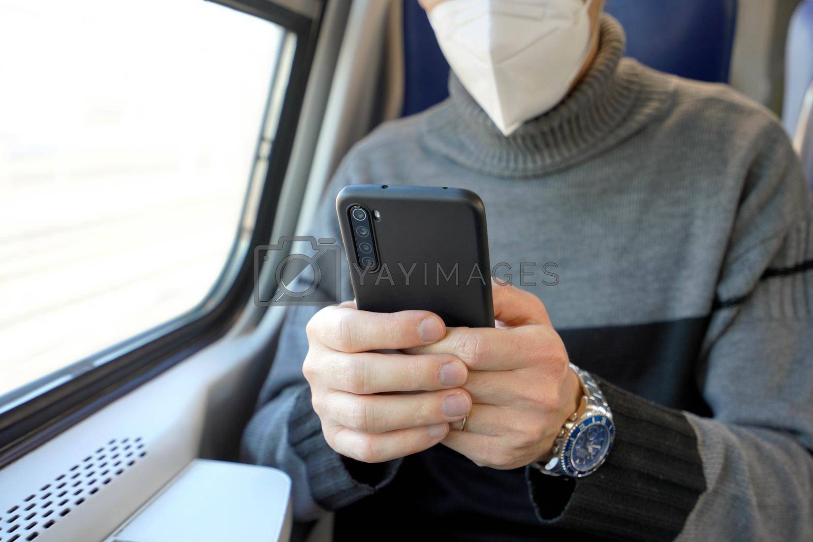 Man on public transport using mobile app wearing medical face mask. Train commuter holding cellphone with mandatory protective mask KN95 FFP2. Focus on the phone.