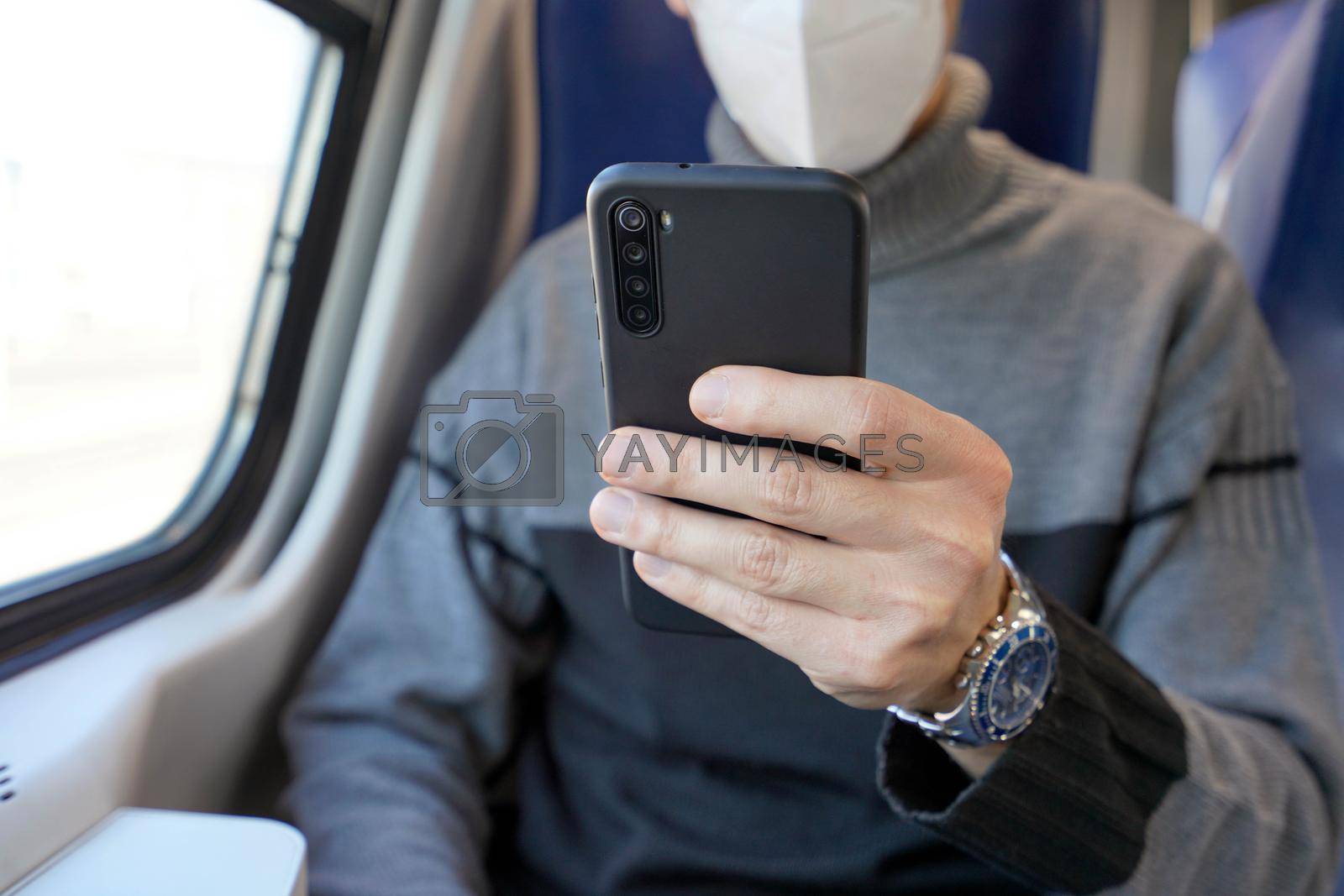 Business man on public transport using mobile app wearing protective face mask. Train passenger holding smartphone covering face with mask KN95 FFP2. Focus on the phone.