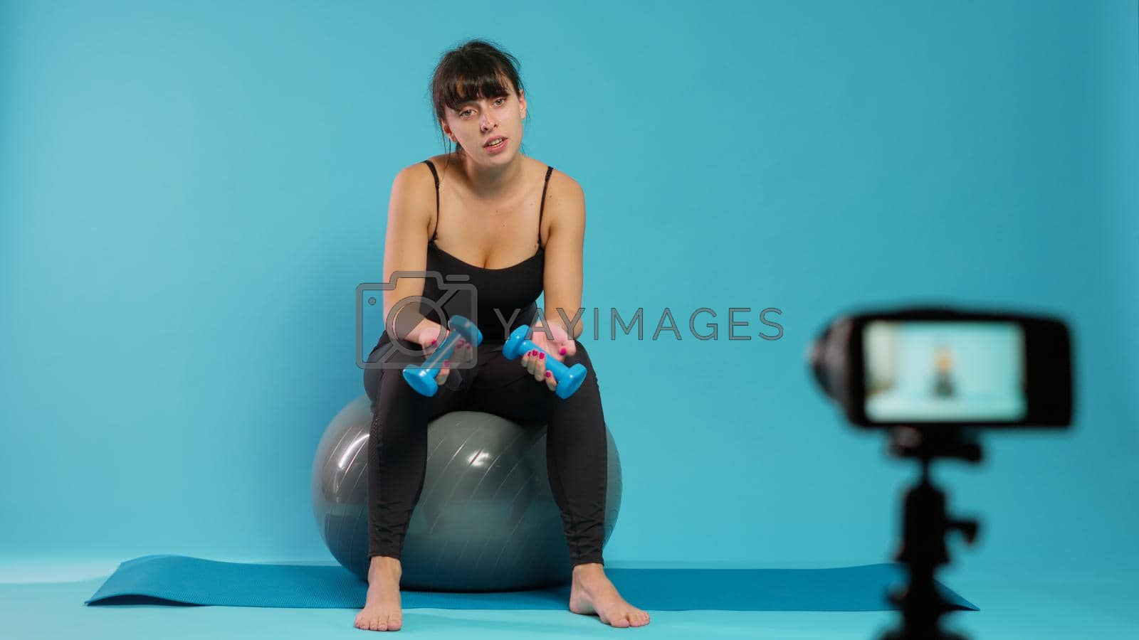 Fitness vlogger recording workout video on camera in studio, using dumbbells to explain lifting technique for sport practice. Muscular woman filming online training lesson on toning ball.