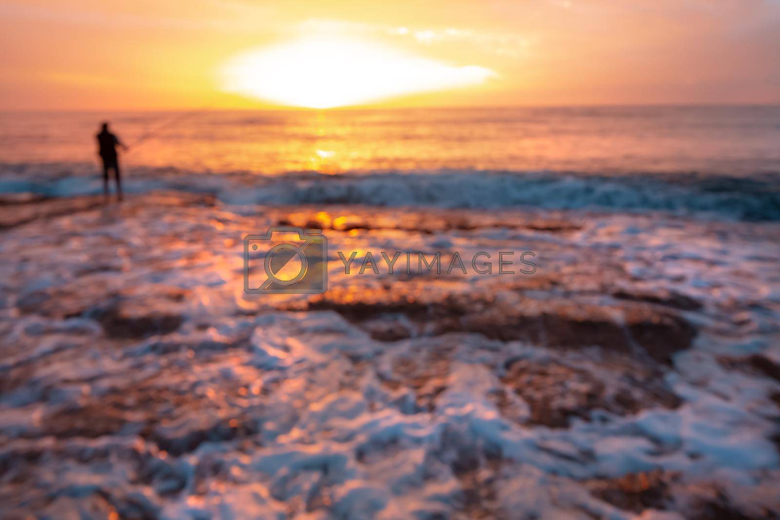Blur Photo of a Sea. Man Fishing over Beautiful Orange Sunset Sky Background. Active Lifestyle on the Beach. Happy Summer Vacation.