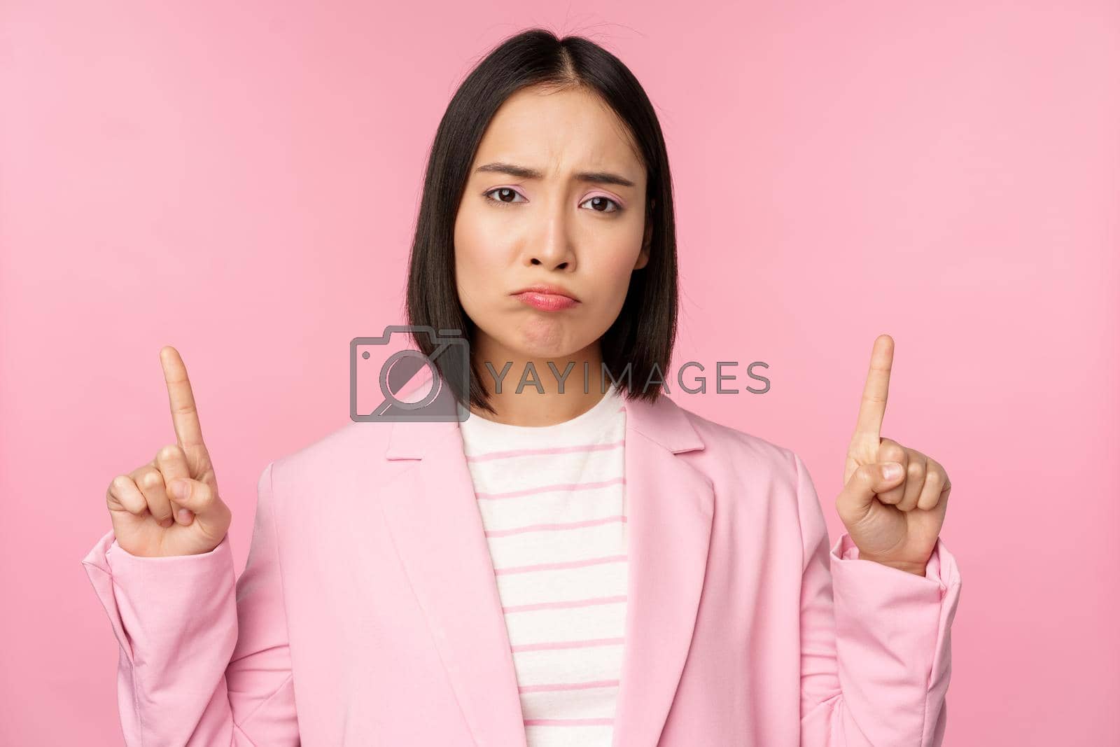 Royalty free image of Sad asian business woman, professional corporate manager pointing fingers up, sulking disappointed, standing over pink background by Benzoix