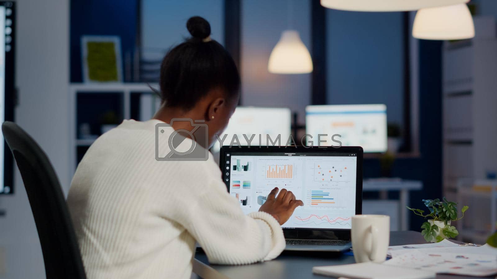 Black woman manager checking business statistics and financial reports on laptop working in start up office late at night. Focused employee doing overtime for job respecting deadline of project