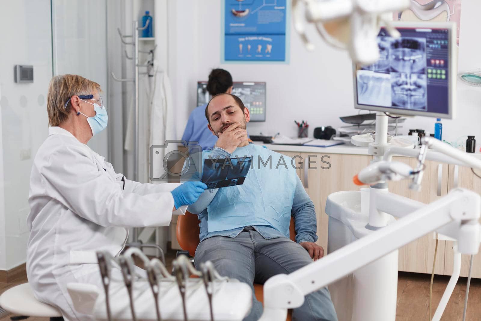 Senior dentist woman with face mask explaining teeth radiography to patient discussing oral treatment to prevent infection during medical examination in dental office. Stomatology concept